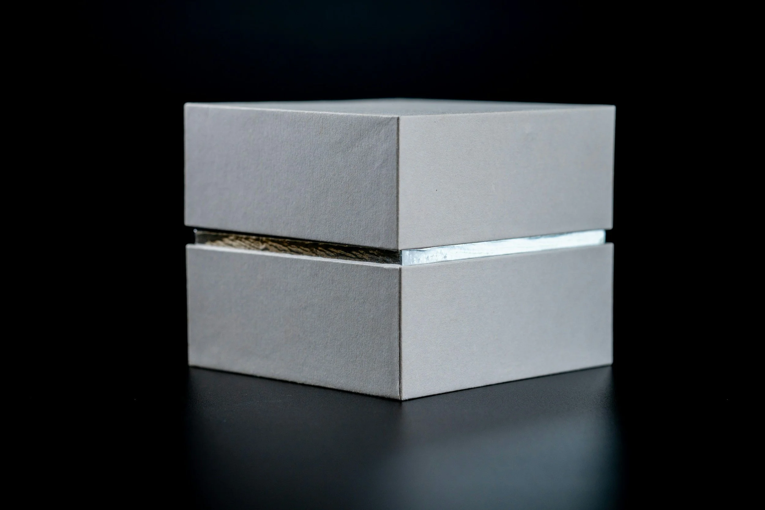 O cutie de carton gri premium, cu gât argintiu pe fundal negru, cu o bandă de material metalic vizibilă între ele. A premium grey cardboard box with a silver neck against a black background, with a strip of metallic material visible between them.