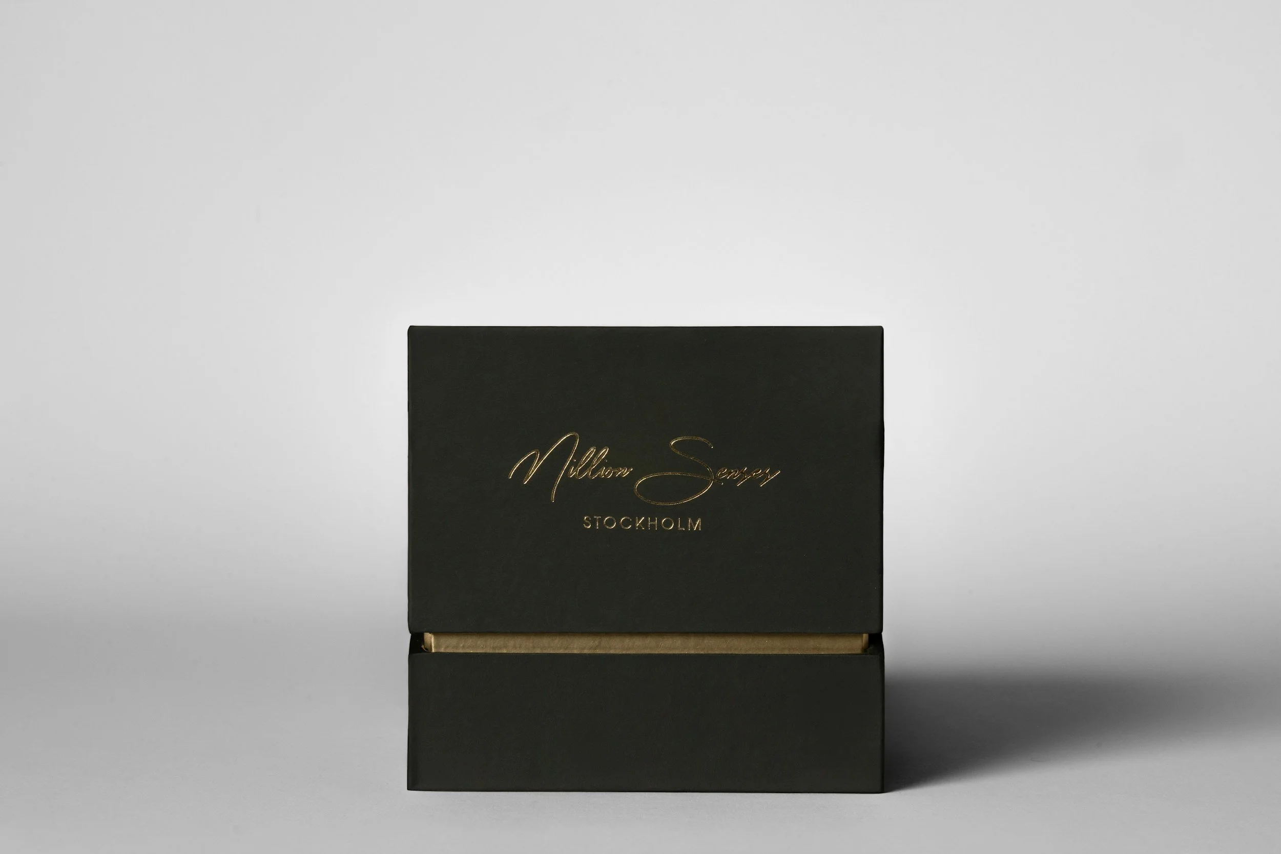 O cutie de bijuterii neagră cu text auriu „Mellom Senap” și „Stockholm” scris cursiv, cu gâta.  A black jewelry box with gold text reading "Mellom Senap" and "Stockholm" in cursive, with a gold-colored neck interior, set against a neutral background.