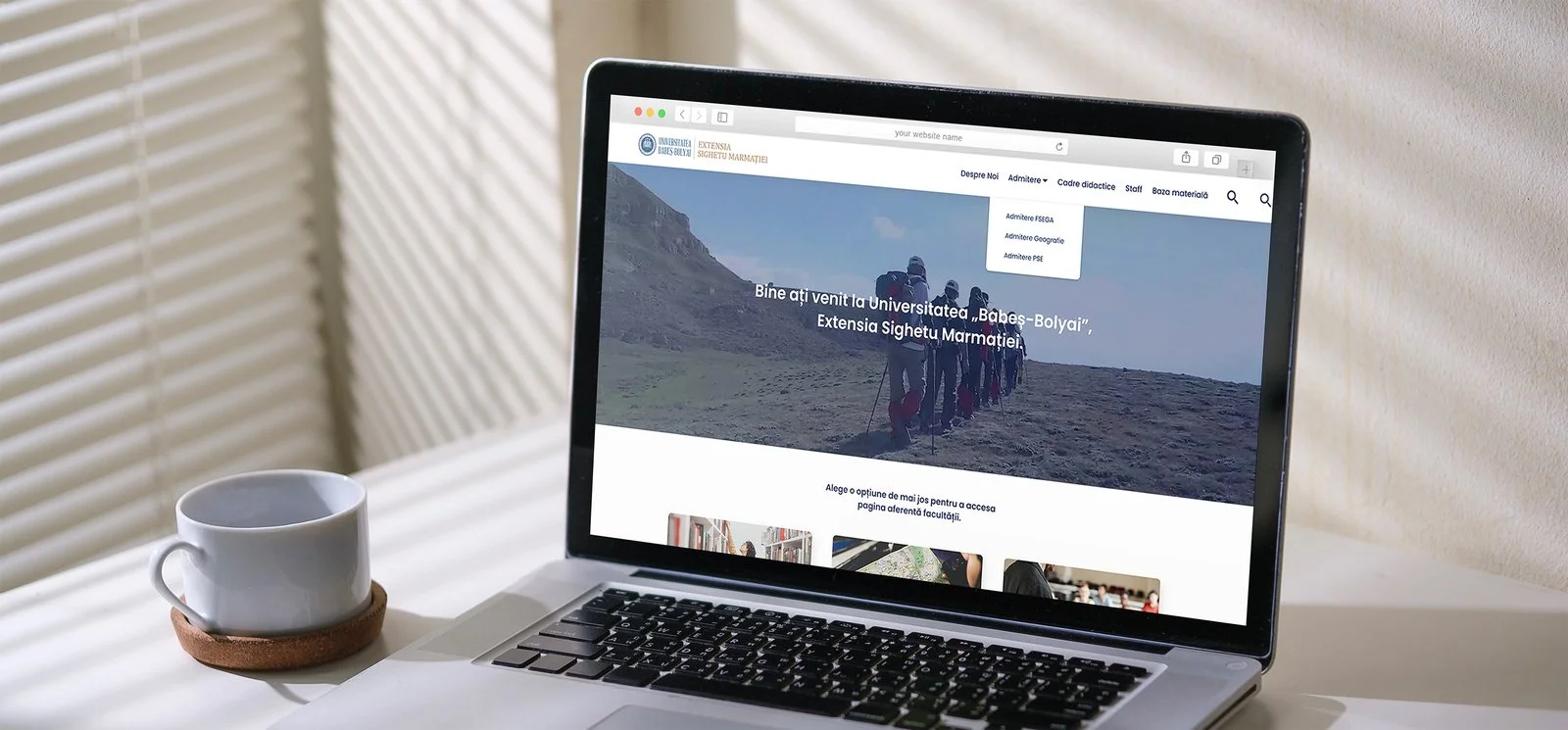A laptop displaying a university website with a mountain landscape and a group of hikers on the screen, next to a white coffee cup on a saucer.