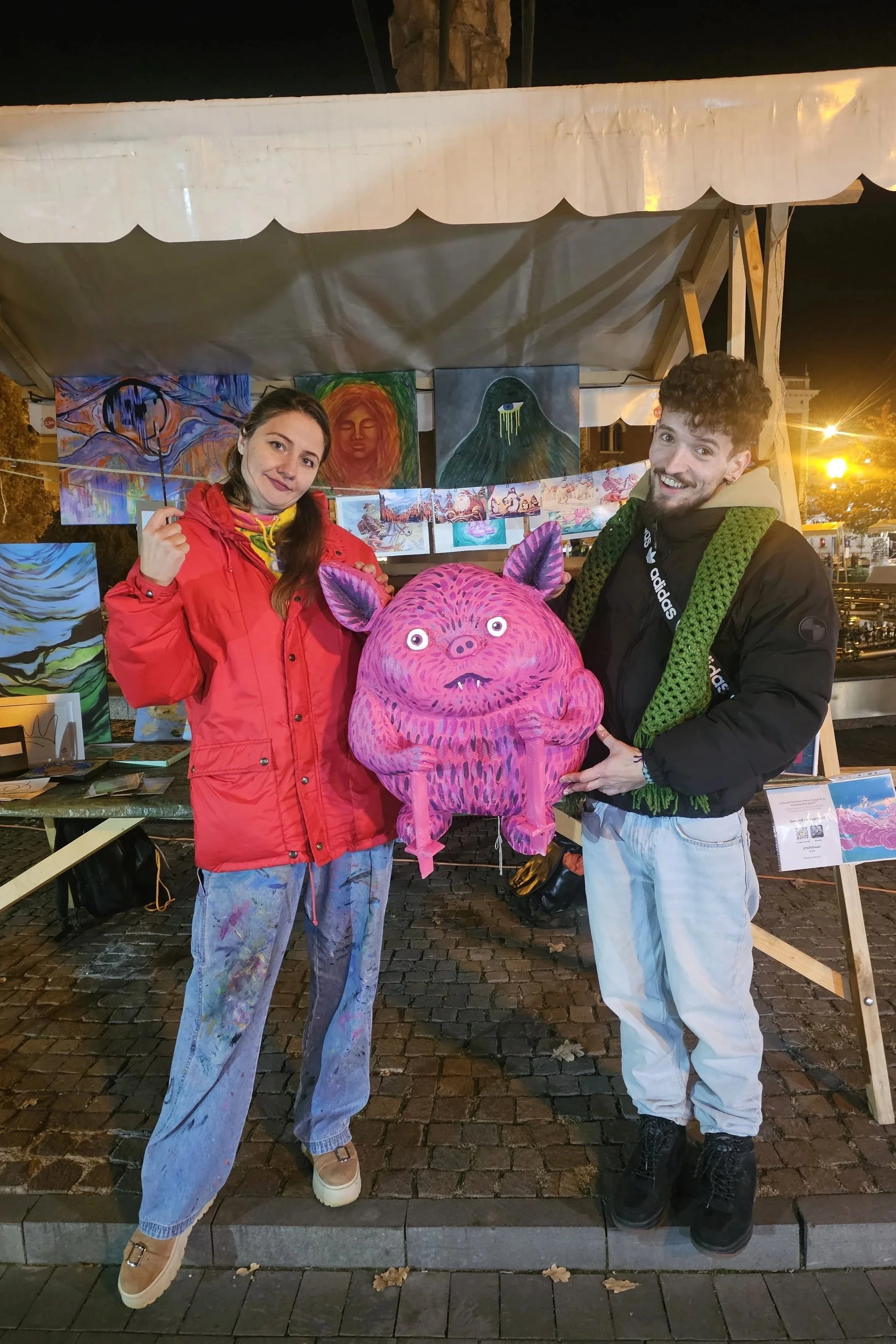 Two people at an art market at night, holding a large, pink, cartoonish creature lantern with cat-like ears and wide eyes. Artwork is displayed behind them, and they are smiling.