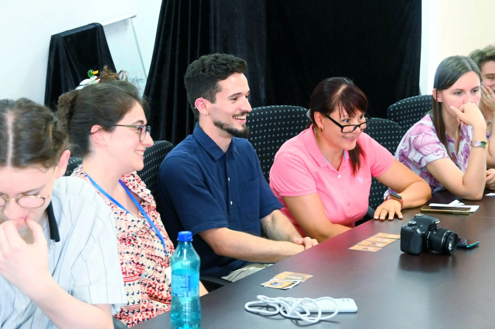 Group of people sitting at a conference table, smiling and engaging in a discussion, with a camera, smartphone, water bottle, and various cards on the table.