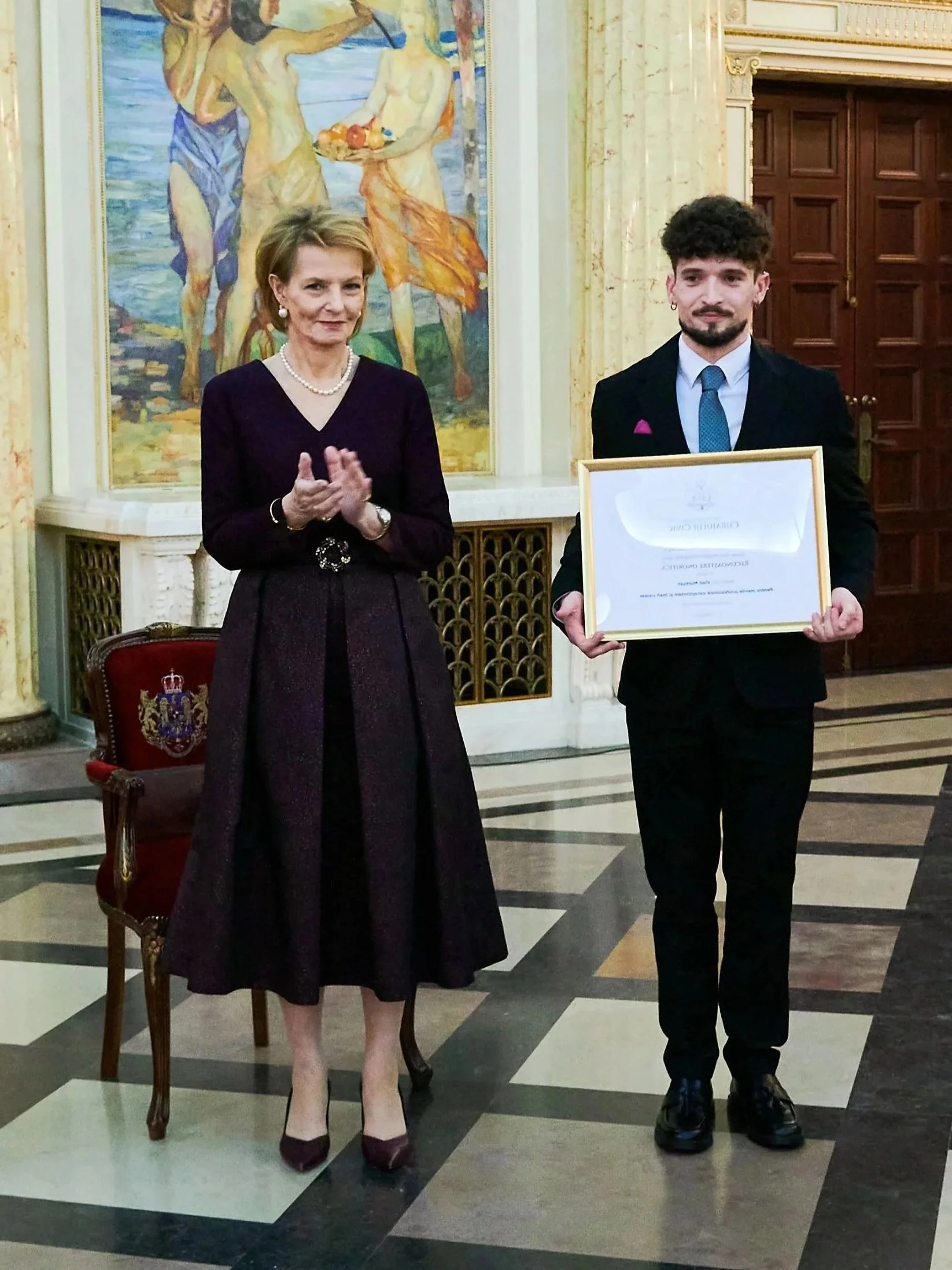 Un tânăr artist primește un certificat, stând lângă o doamnă care aplaudă (posibil Principesa Margareta a României), în timpul unei ceremonii oficiale într-o sală regală.