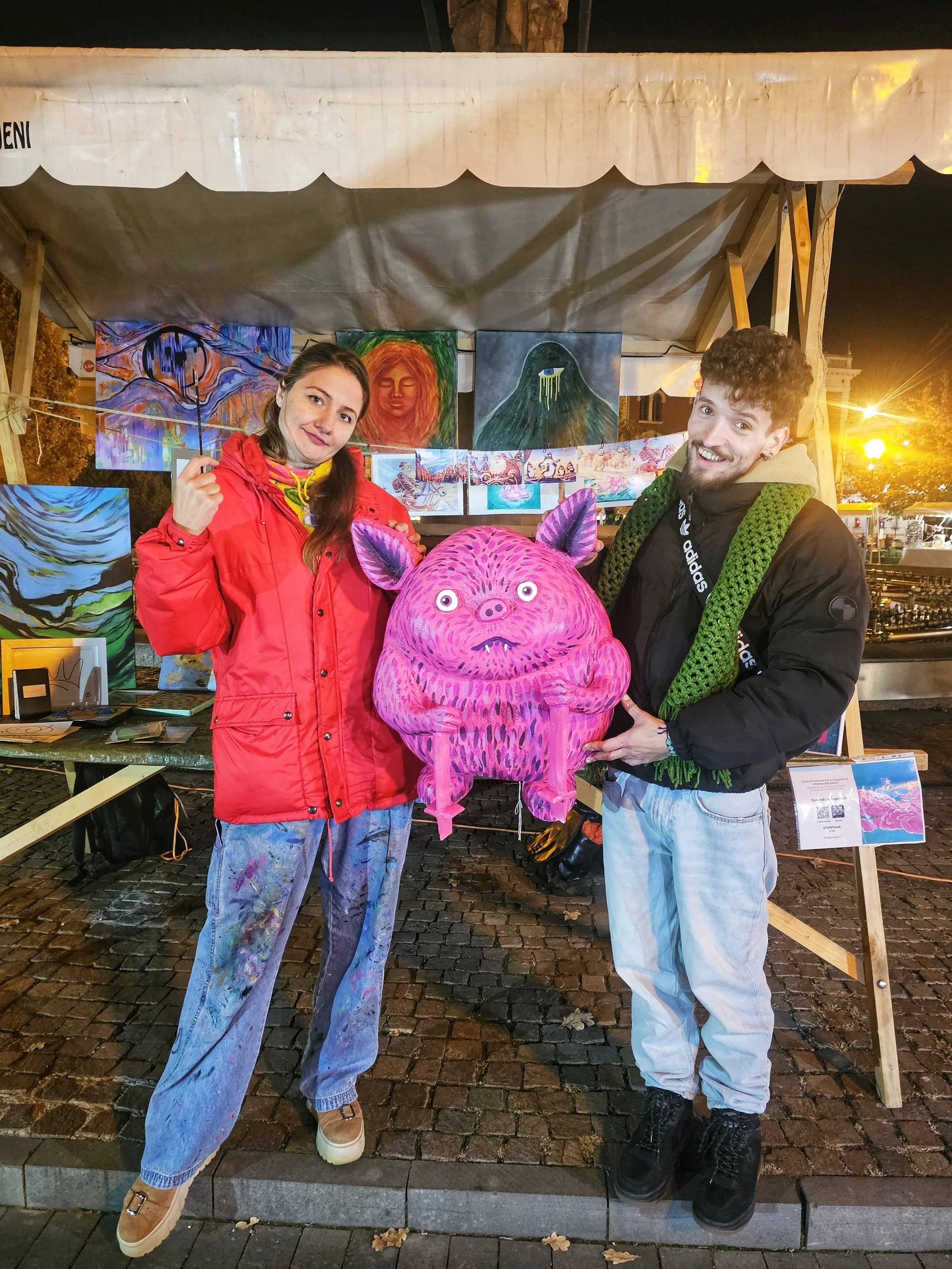 Two people standing in front of a booth displaying colorful artwork at night. They are holding a pink, cartoonish creature with large eyes and pointed ears. The woman on the left is wearing a red jacket and has long brown hair, while the man on the right has curly hair, a black jacket with green scarf, and is smiling.
