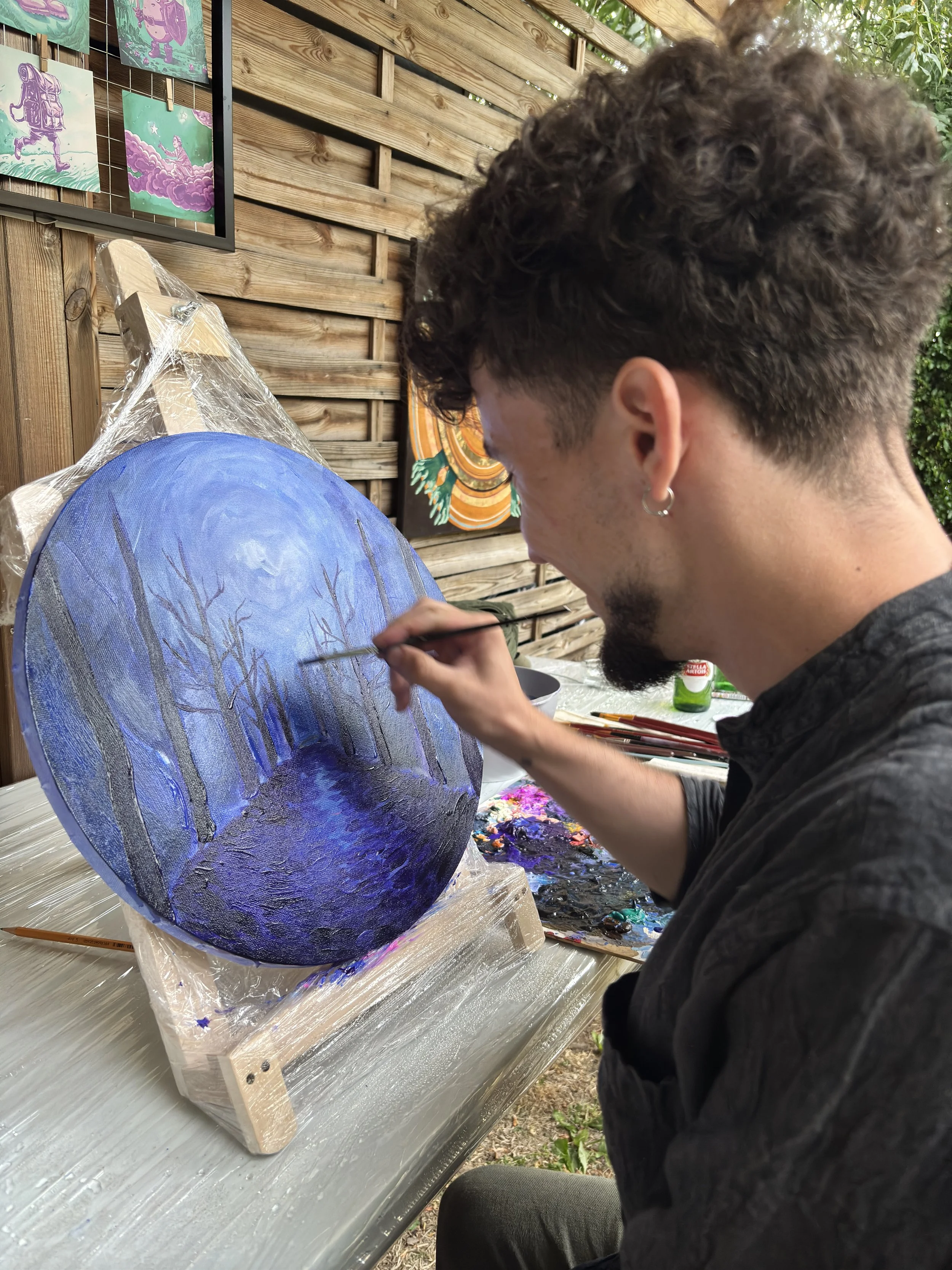 A man with curly hair and earrings painting a moonlit forest scene on a round canvas in an outdoor art studio. The scene depicts tall trees and a glowing moon. Painting supplies and photos of artwork are visible in the background.