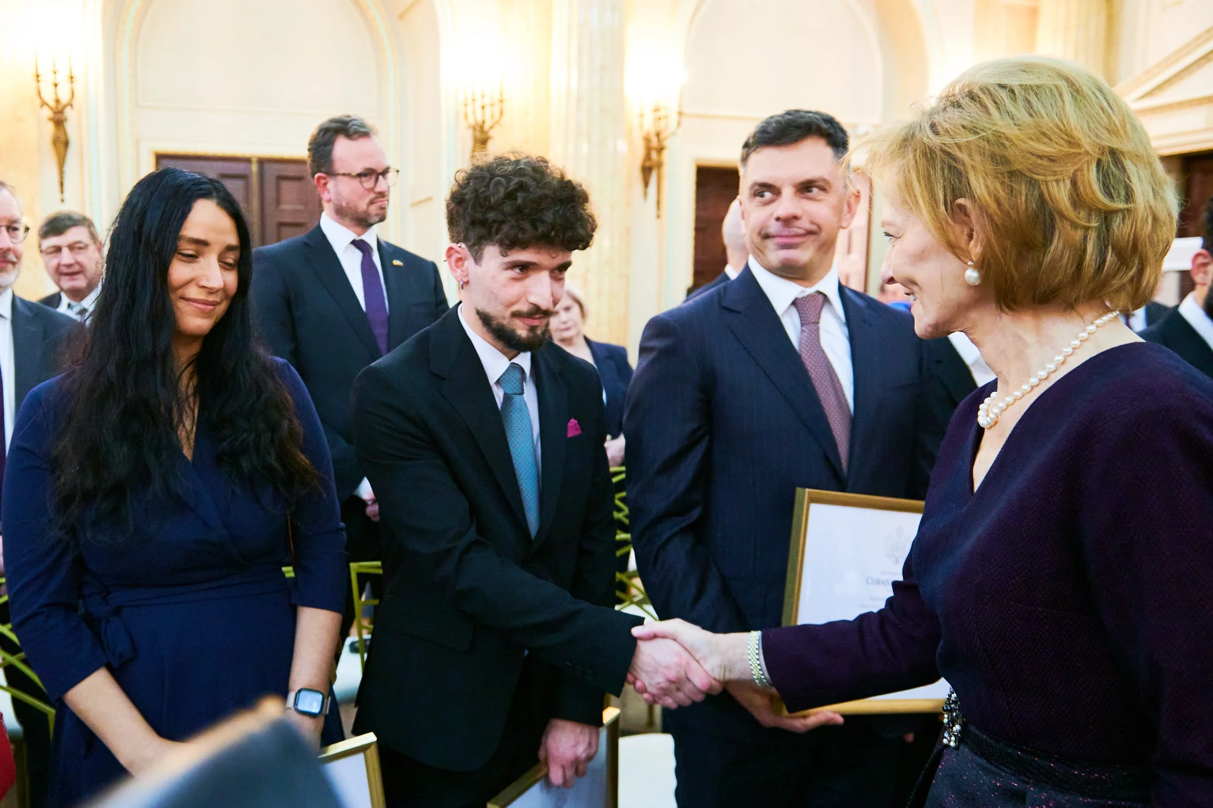 O femeie în rochie violet și colier de perle dă mâna cu un bărbat în costum la un eveniment formal, în timp ce alți invitați privesc.