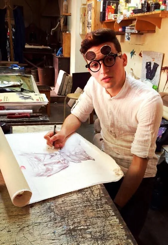 Young man sitting at a worn wooden table in an art studio, working on a sketch or drawing on large paper, wearing glasses and a light-colored shirt.