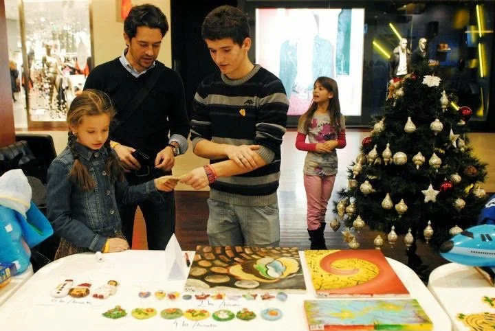 A family of four shopping or browsing holiday-themed arts and crafts at a table in a mall, with a decorated Christmas tree nearby.
