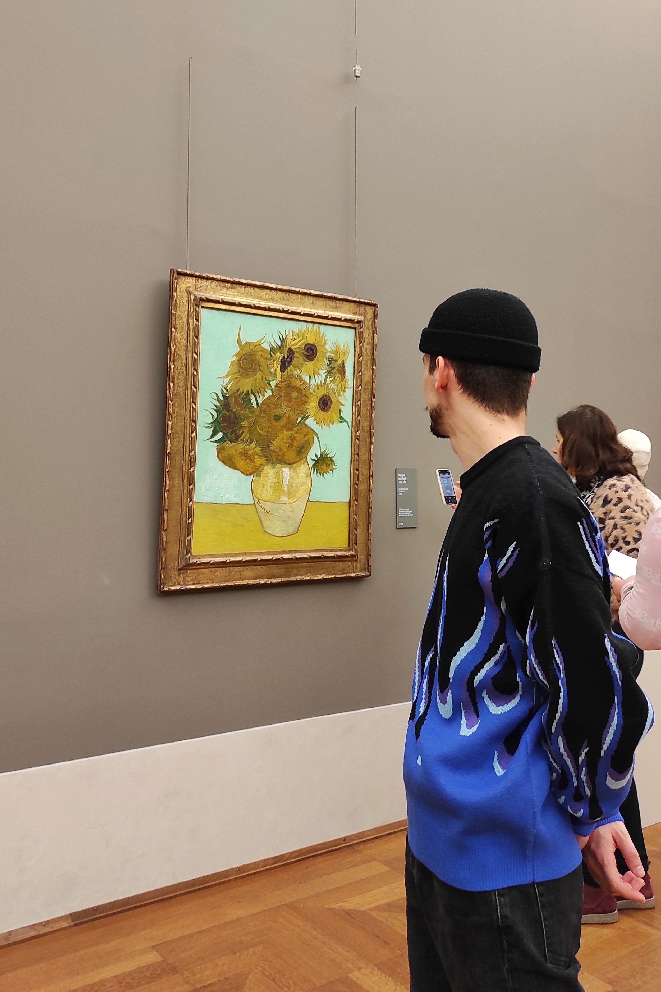 A man in a black beanie and blue and black sweater observing a painting of sunflowers in a gold frame, displayed on a gray wall at an art gallery.