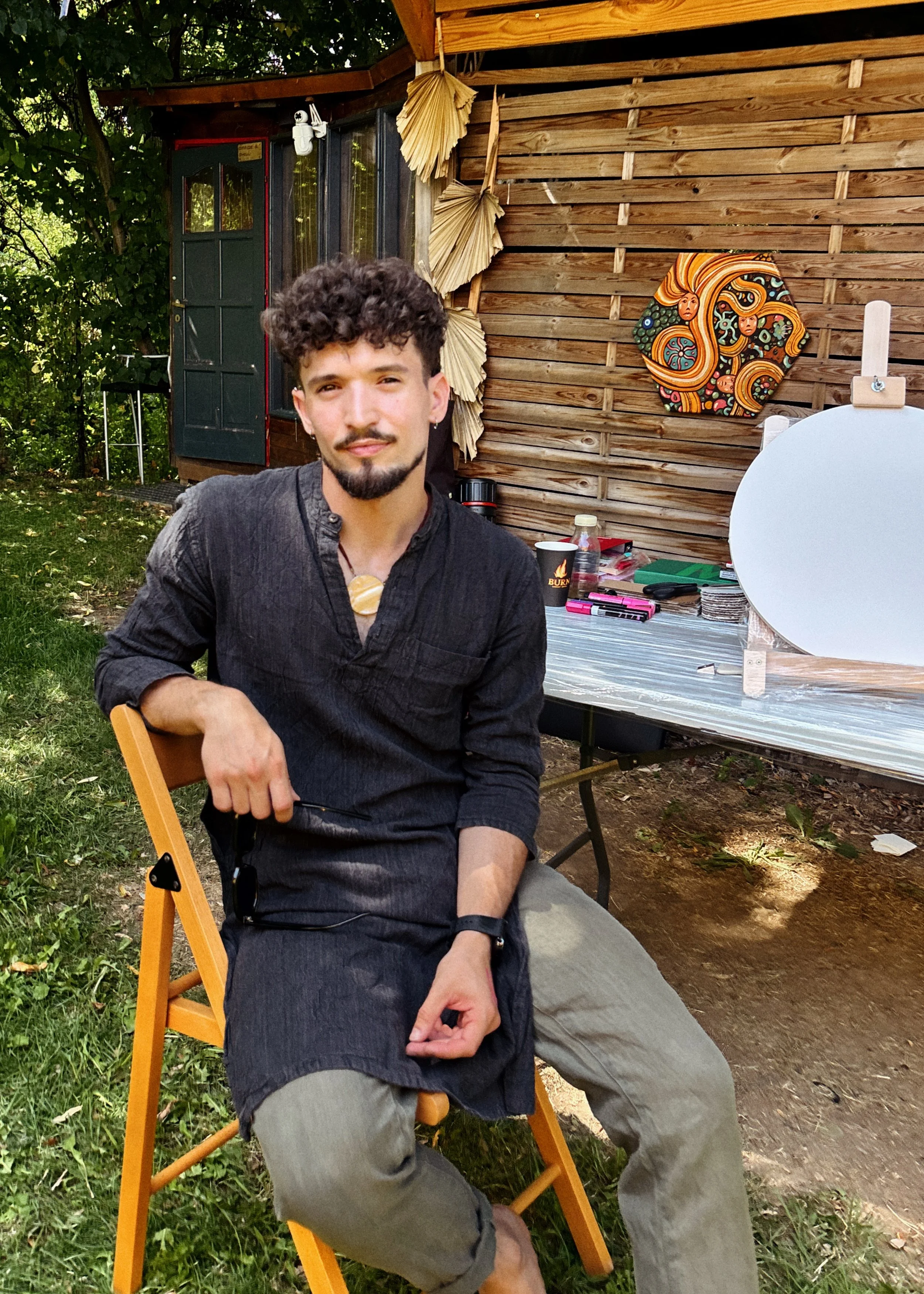 A man with dark curly hair, a beard, and earrings, sitting on a wooden chair outdoors, wearing a dark long-sleeve shirt and light pants. Behind him is a wooden structure, part of which holds a colorful piece of artwork, with a table holding various items including a paper reflector and other small objects.