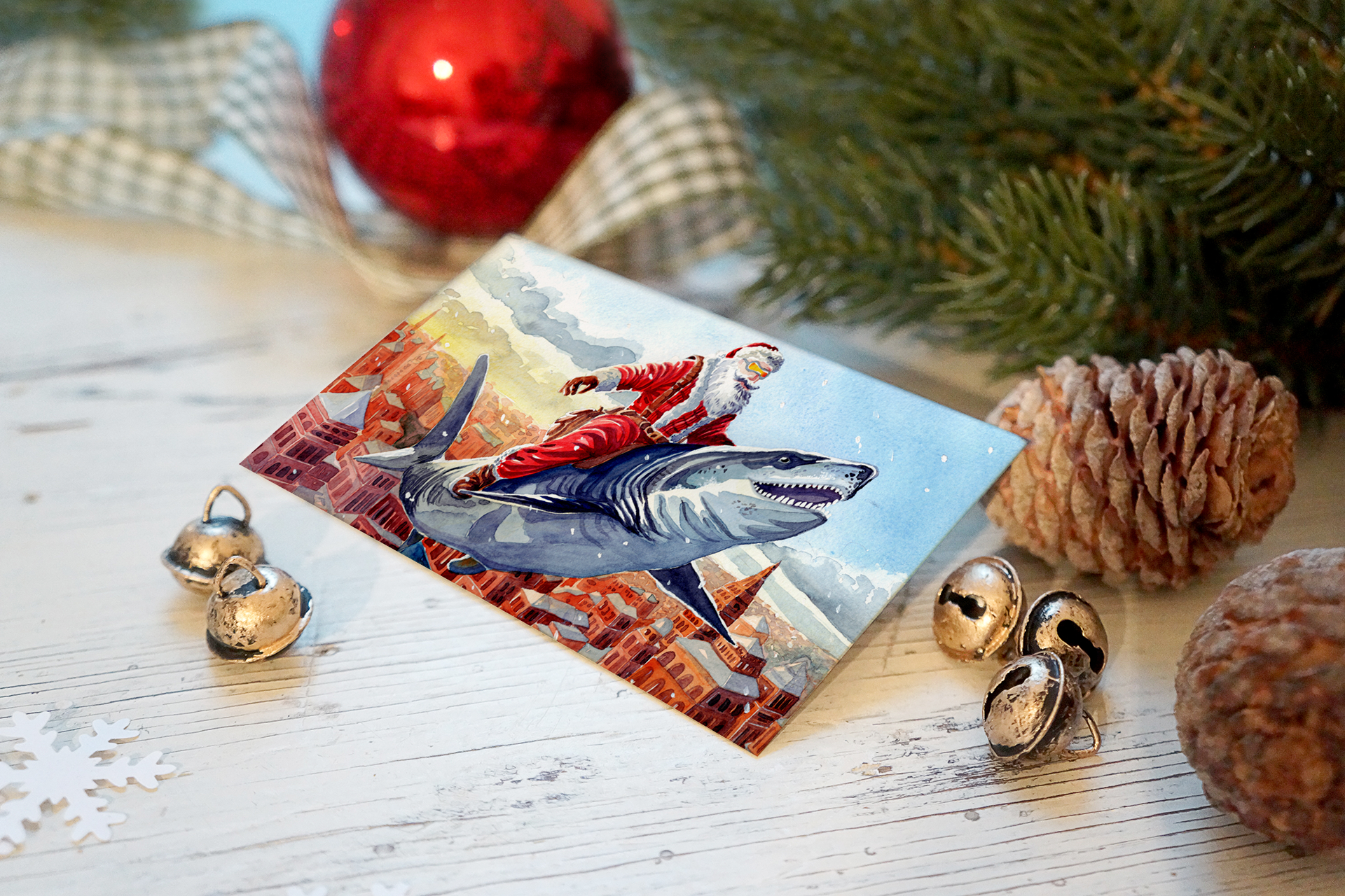 Christmas greeting card depicting Santa Claus riding a large shark over a cityscape with mountains in the background, surrounded by holiday decorations including silver jingle bells, pinecones, a red ornament, an evergreen branch, and a snowflake.