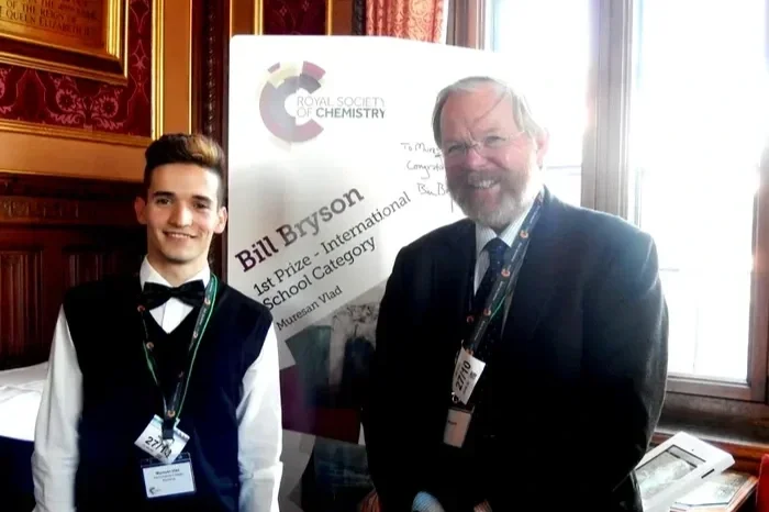 Un tânăr și un bărbat mai în vârstă stând înăuntru, zâmbind pentru cameră. În spatele lor se află un afiș al Societății Regale de Chimie, cu numele Bill Bryson și indicând un premiu întâi la o categorie școlară internațională.