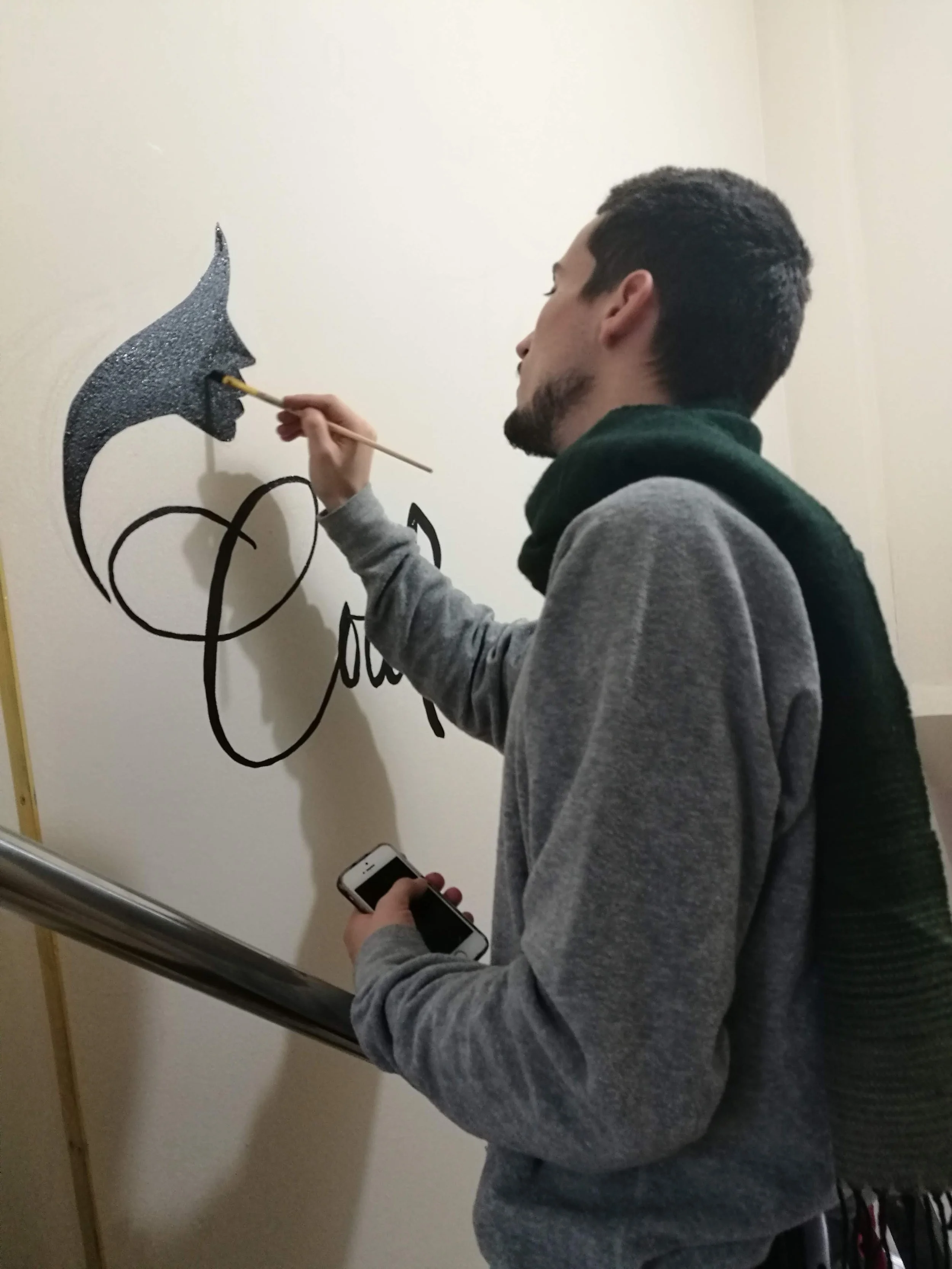 Un bărbat pictează o față neagră strălucitoare pe un perete alb dintr-un salon de înfrumusețare, ținând un smartphone în mâna stângă.  A man painting a glittery black face on a white beauty studio wall, holding a smartphone in his left hand.