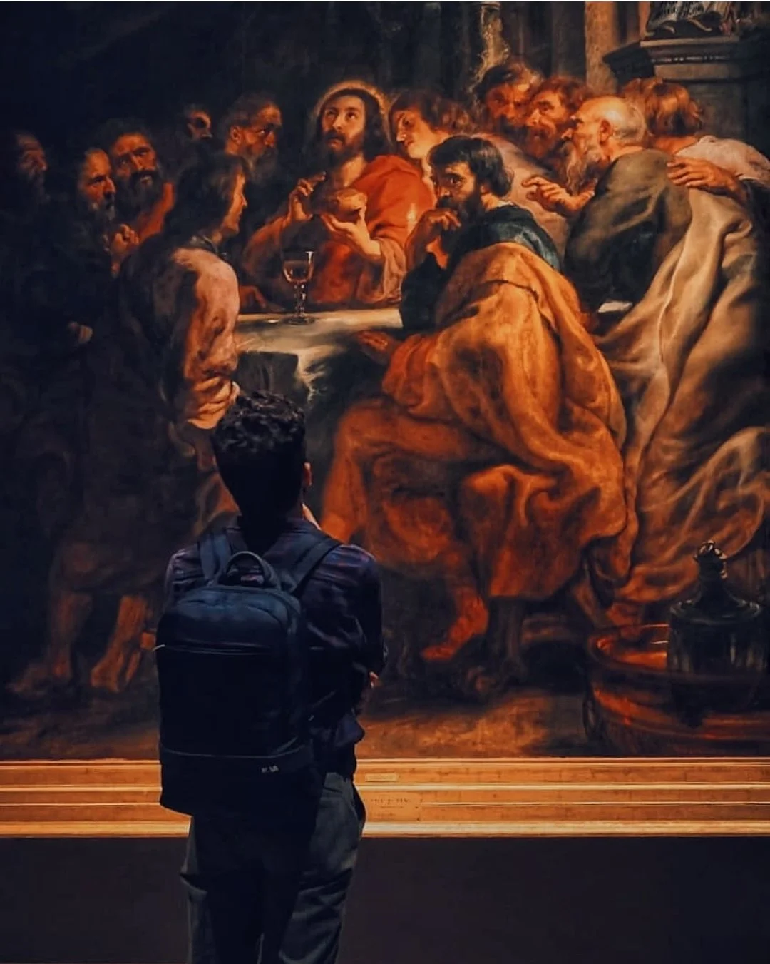 A person with a backpack is observing a large painting of the Last Supper in a museum.