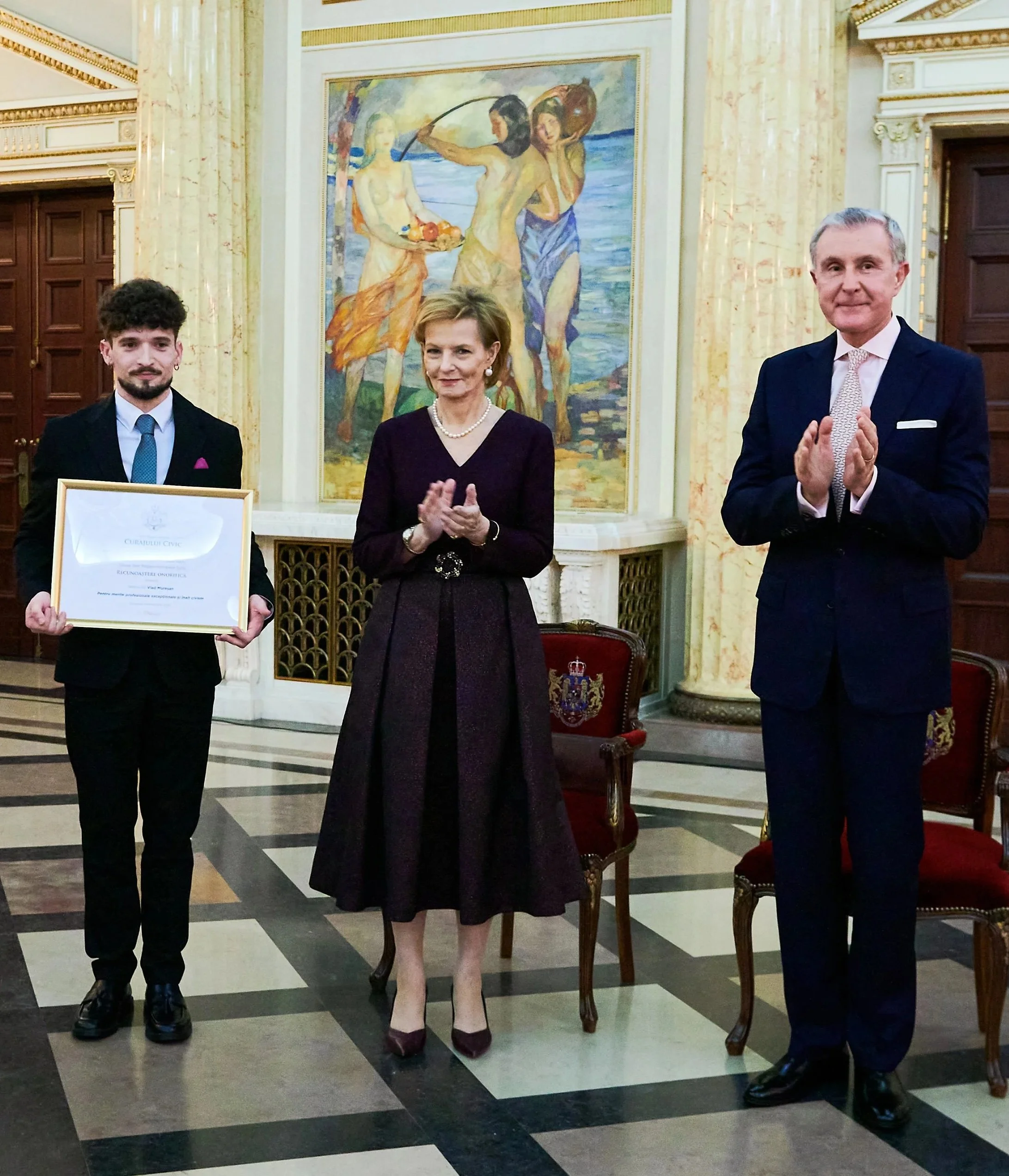 Un tânăr primind un premiu, stând lângă o femeie și un bărbat în ținută elegantă într-o cameră grandioasă, cu un tablou mare reprezentând personaje lângă o întindere de apă în fundal.