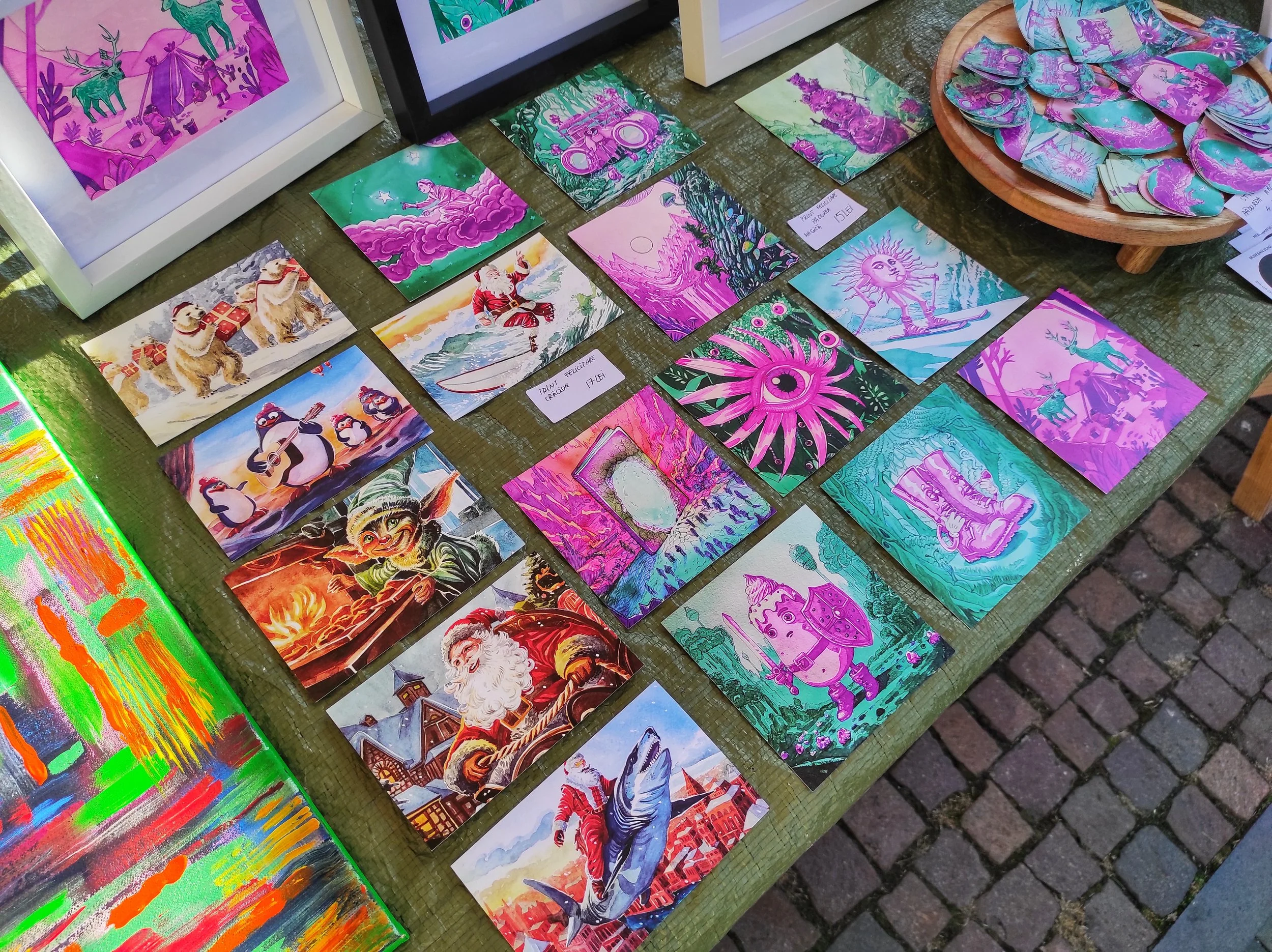 Opere de artă și ilustrații colorate, răspândite pe o masă verde, cu teme fantastice, nautice, de sărbători și fanteziste, cu un bol cu ​​pietre pictate care prezintă opere de artă similare la un târg de artă.