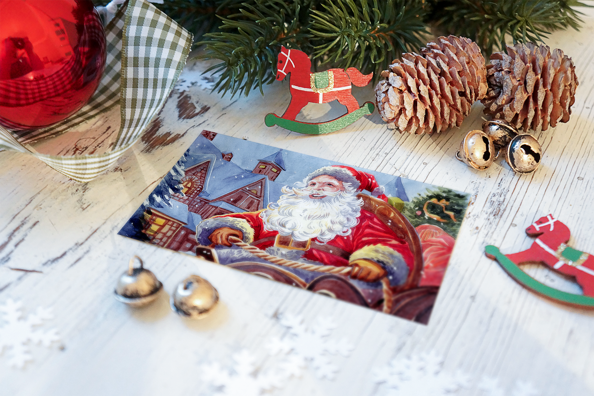 Christmas-themed decorations including Santa Claus illustration, pinecones, jingle bells, a red ornament, and a Santa rocking horse on a white wooden surface with pine branches and checkered cloths.