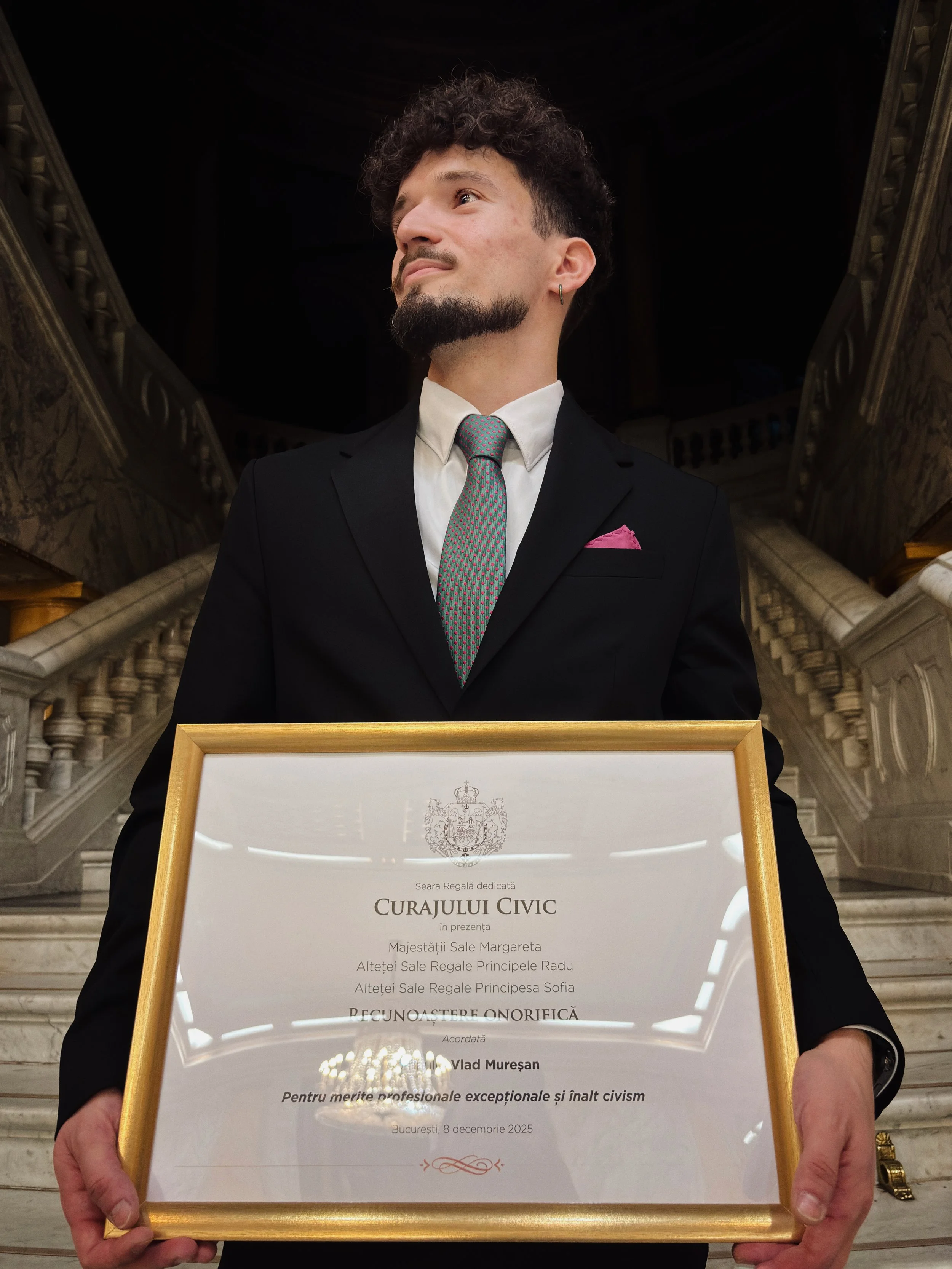 Bărbat în costum negru ținând un certificat înrămat în interiorul unei clădiri cu scara impunătoare.