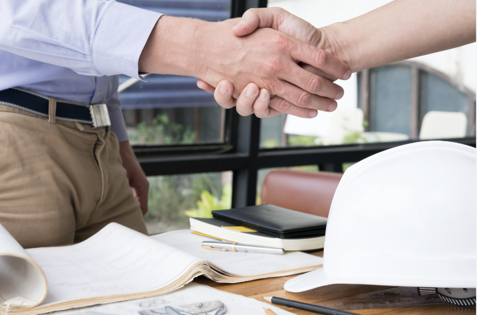 Two people shaking hands over a table with documents, a white safety helmet, and notebooks in an office setting.