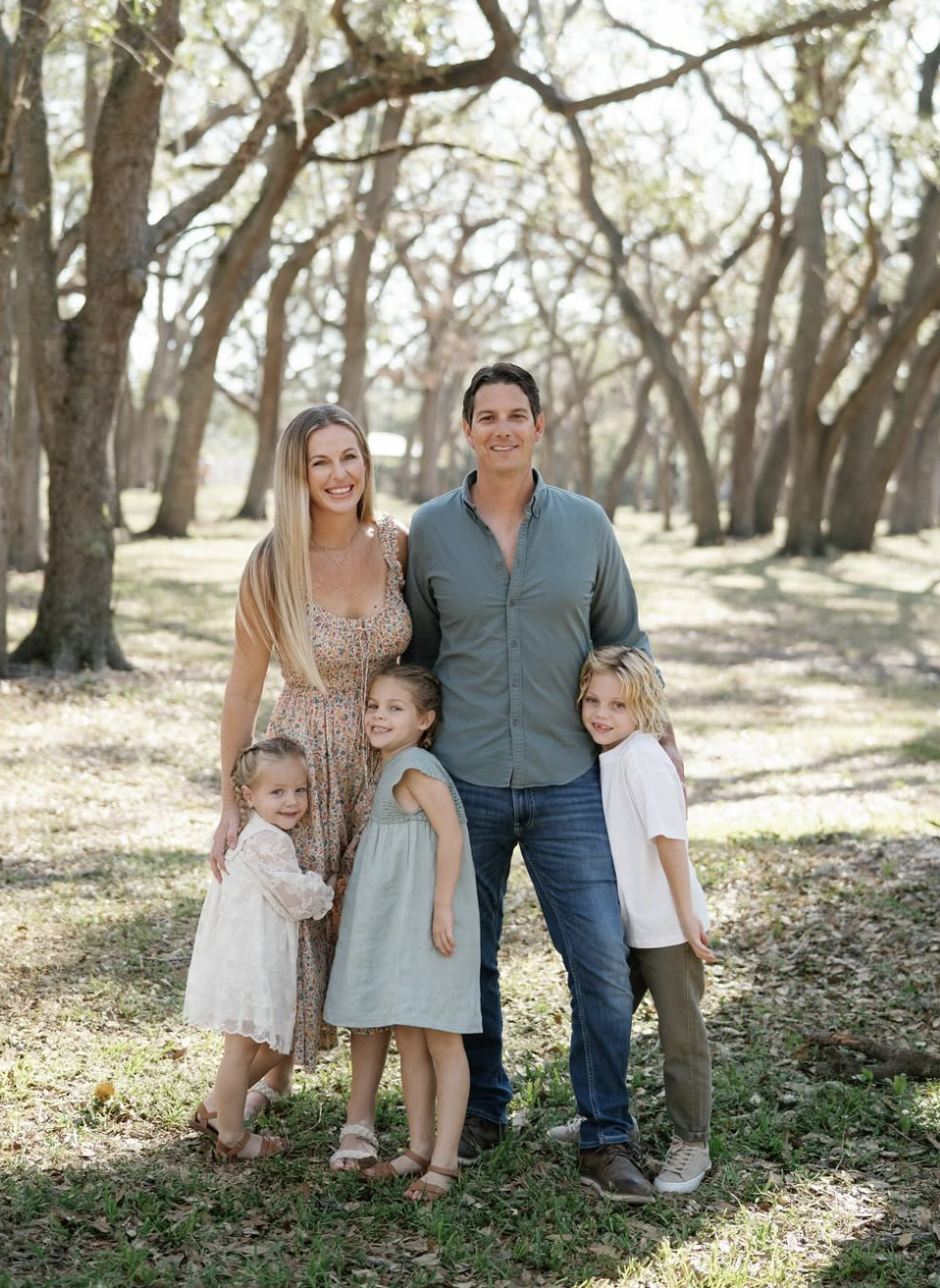 A family of five posing outdoors in a wooded area with sunlight filtering through trees. The family includes a woman, a man, and three young girls, all standing close together and smiling at the camera.