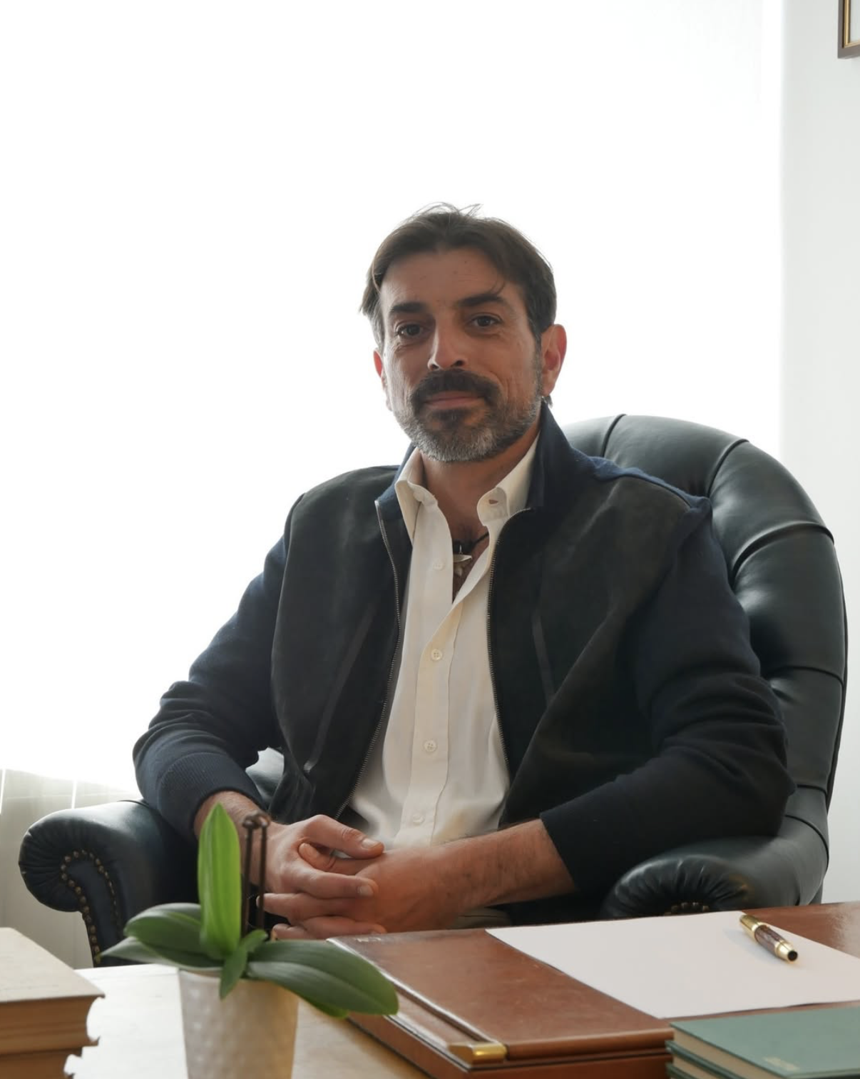Uomo seduto in una stanza, con scrivania e penna, in posa professionale.