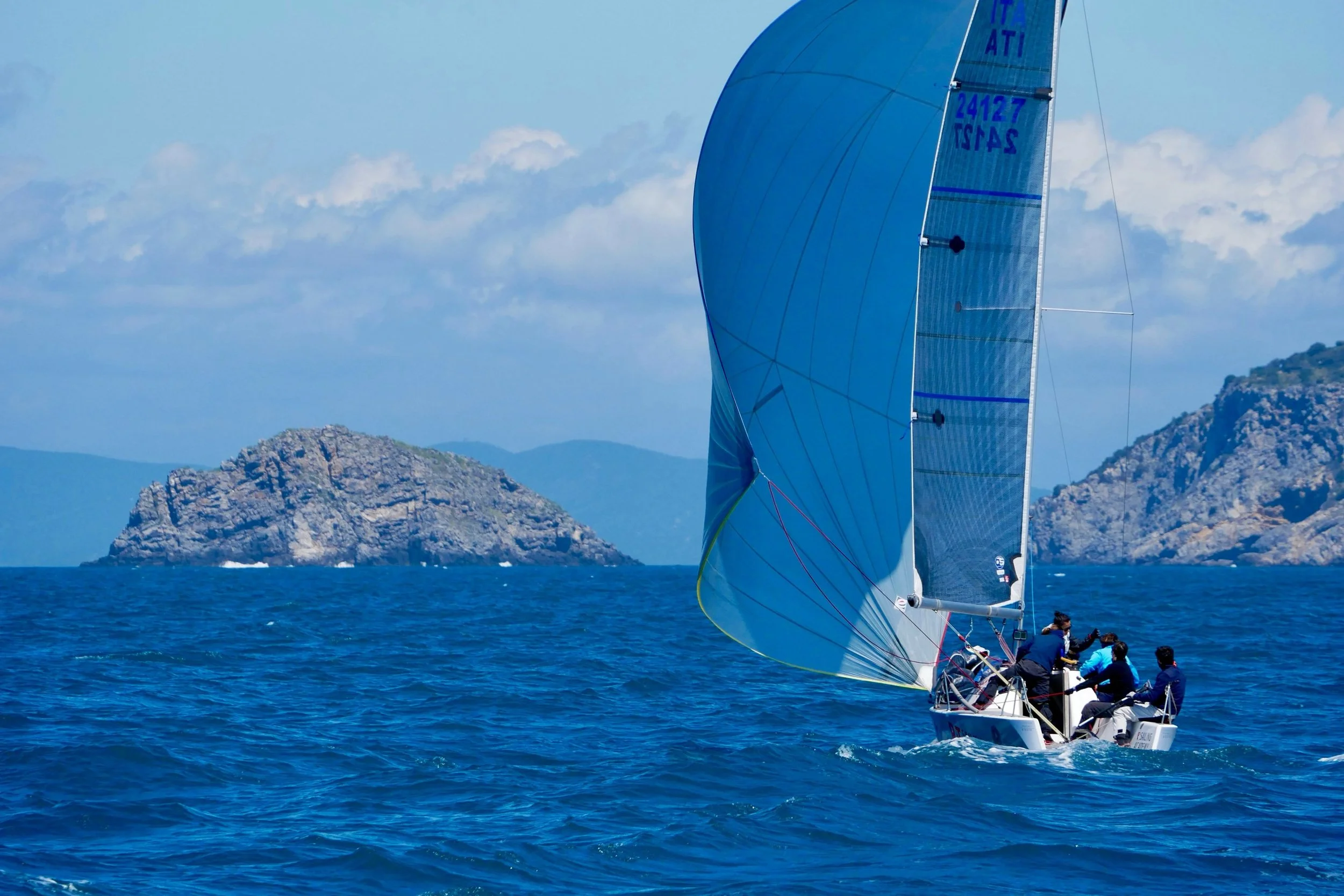 Una barca a vela con una grande vela blu naviga nel mare con isole rocciose sullo sfondo.