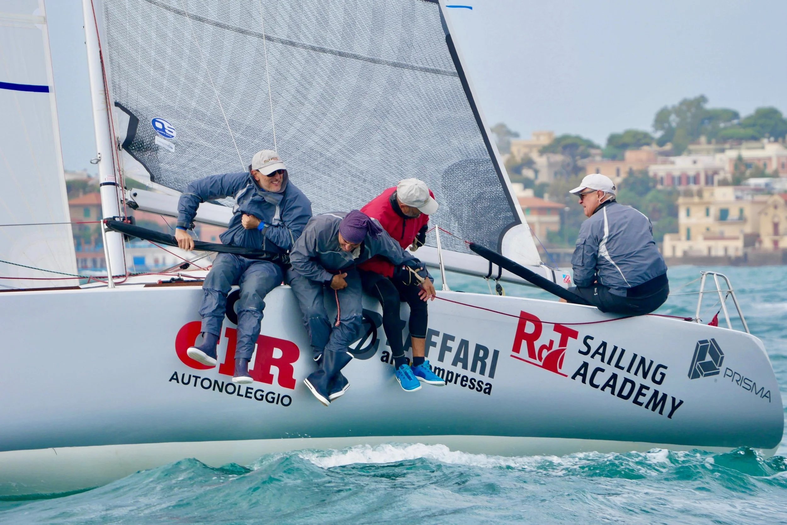 Gruppo di persone in equipaggiamento da regata su una barca a vela, con una città sullo sfondo, che sembrano collaborare per mantenere l'equilibrio della barca durante una regata.