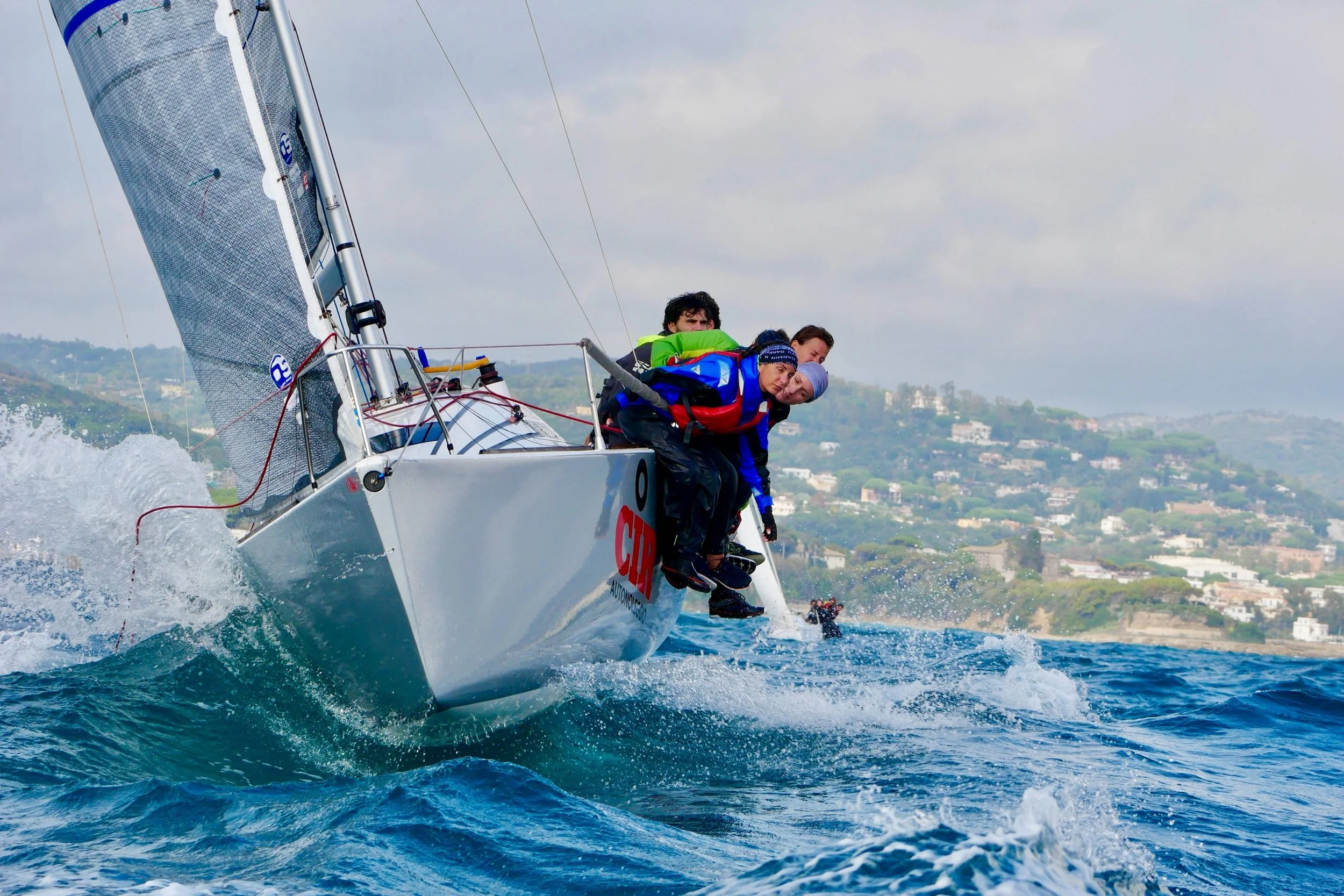 Un equipaggio di tre persone si trova sul ponte di una barca a vela che naviga in mare, con le persone inclinate verso il mare per stabilizzare la barca.