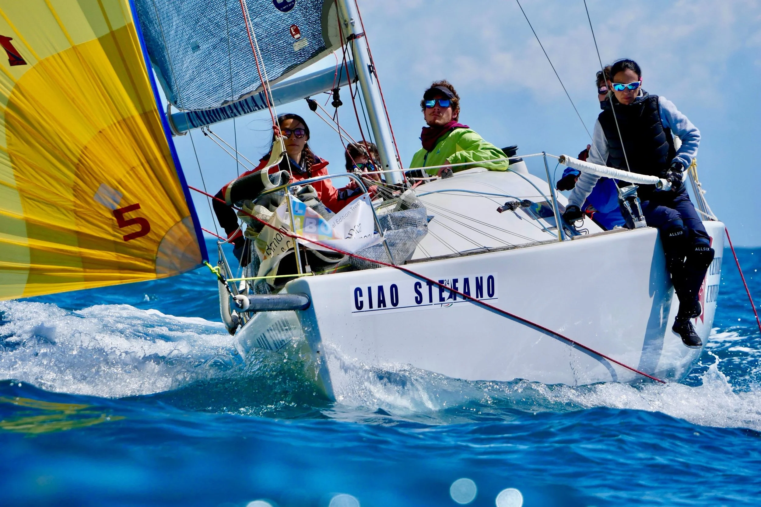 Gruppo di persone su una barca a vela in mare, con vele colorate e il nome 'CIAO STEFANO' visibile sul lato della barca.