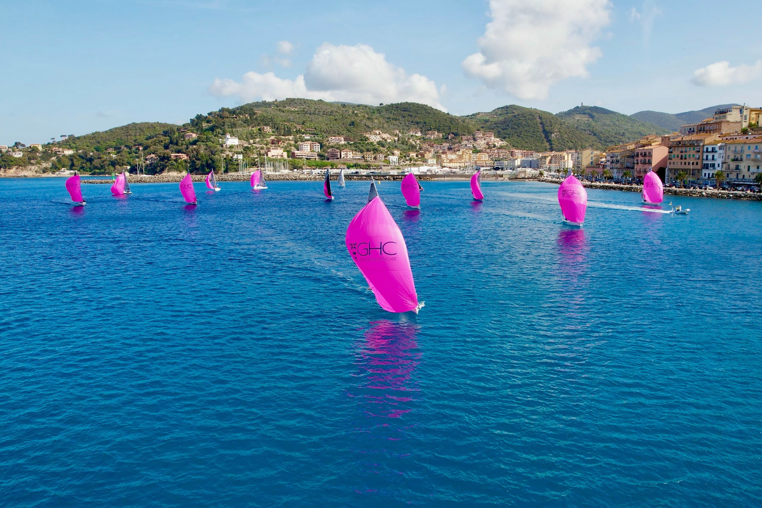 Barche a vela con vele rosa che navigano in mare davanti a una città costiera e colline verdi, con cielo sereno e nuvole