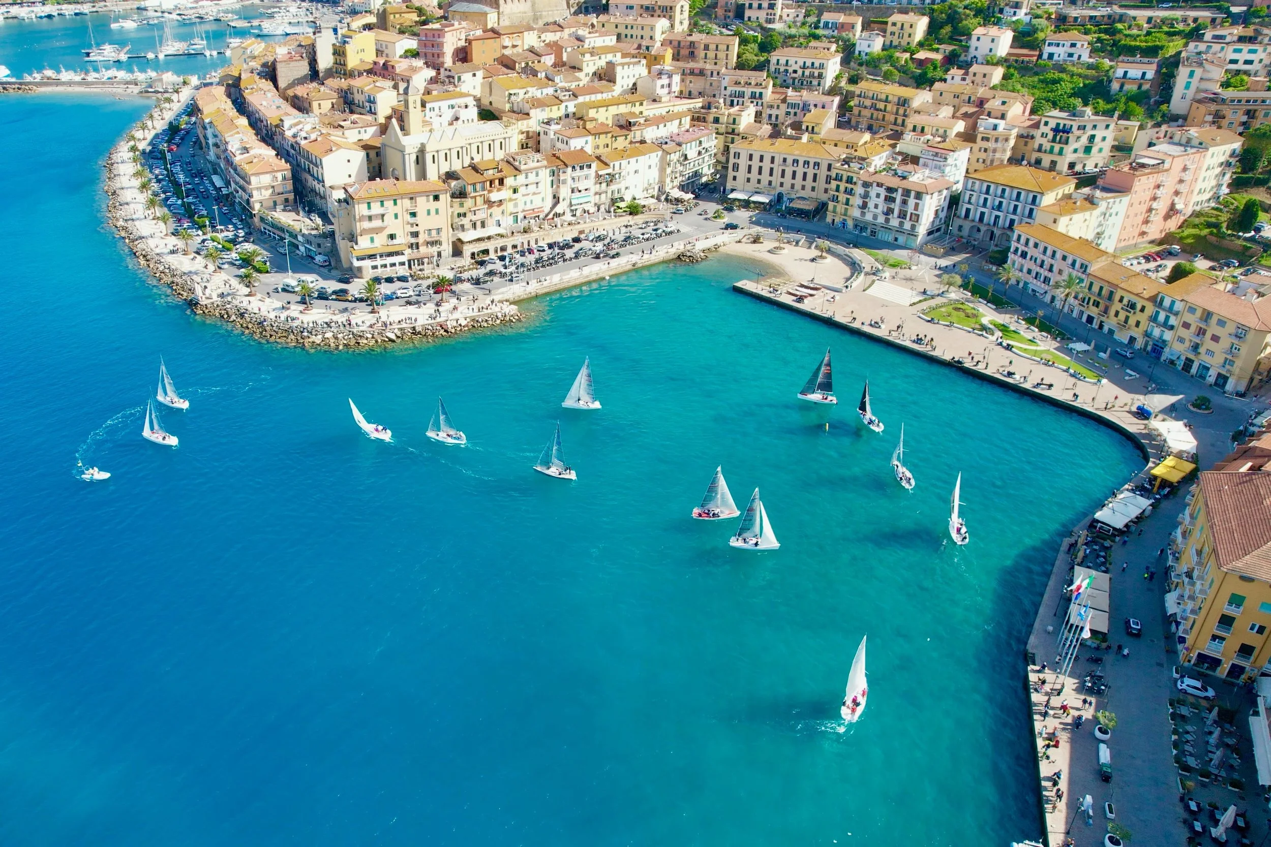 Veduta aerea di un porto con varie barche a vela che navigano l'acqua turchese, un lungomare affollato con edifici colorati e alcuni ristoranti, e un'area pedonale con persone che camminano lungo il mare.