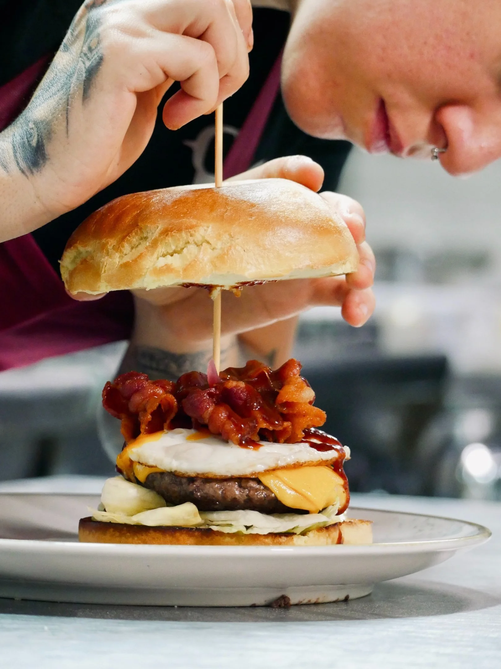 Persona che prepara un hamburger con carne, formaggio, bacon, insalata, cipolla e salsa, appoggiato su un piatto bianco.