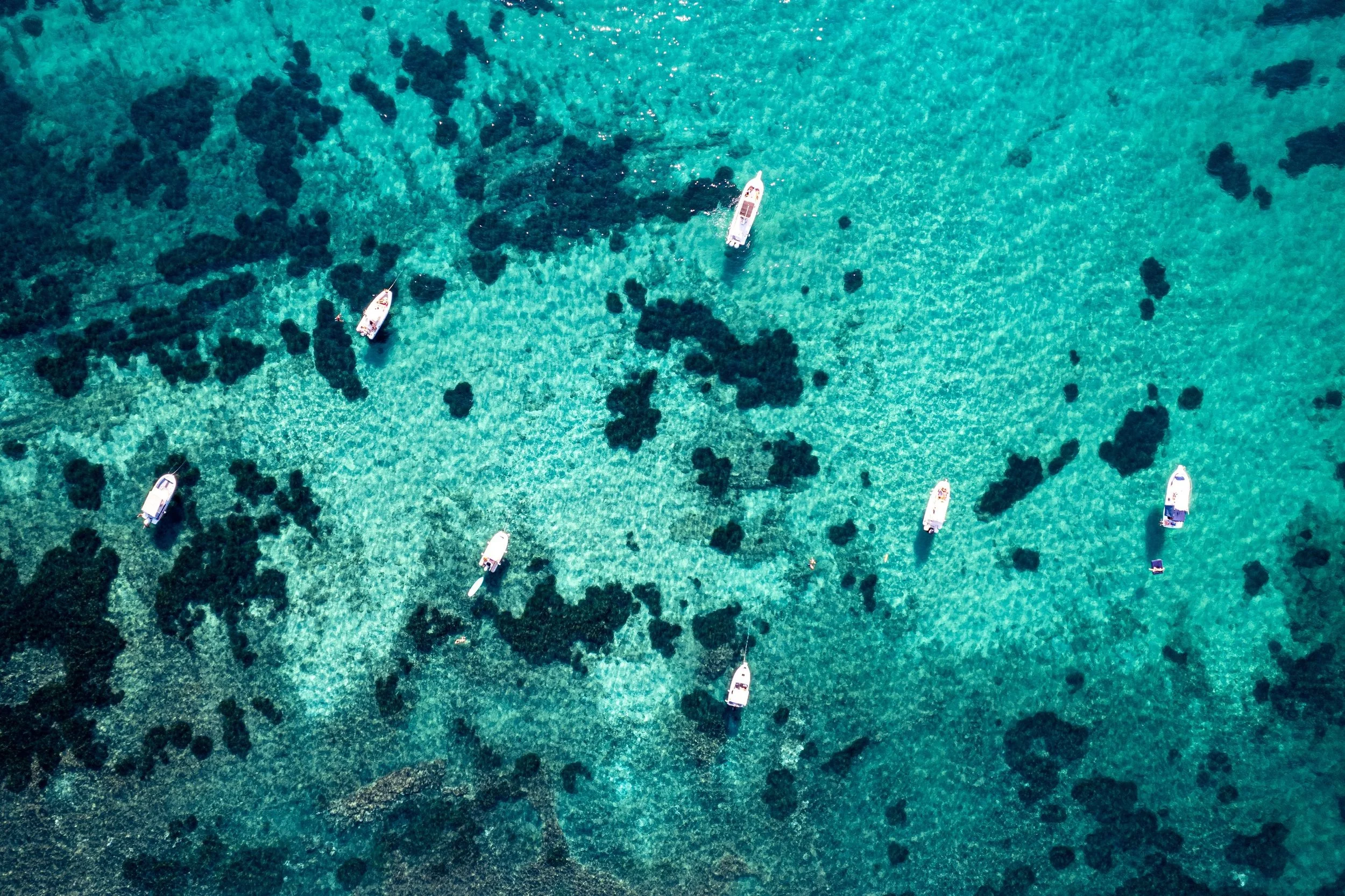 Immagine aerea di barche ormeggiate in acque cristalline, con scogli e fondale visibile sotto l'acqua.