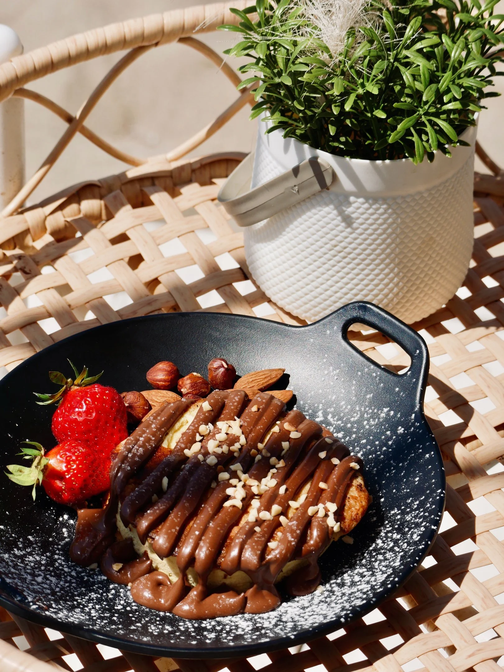 Dolce con cioccolato, fragole, nocciole e mandorle su un piatto nero su una seduta di vimini, con una pianta in una pianta bianca in sfondo.