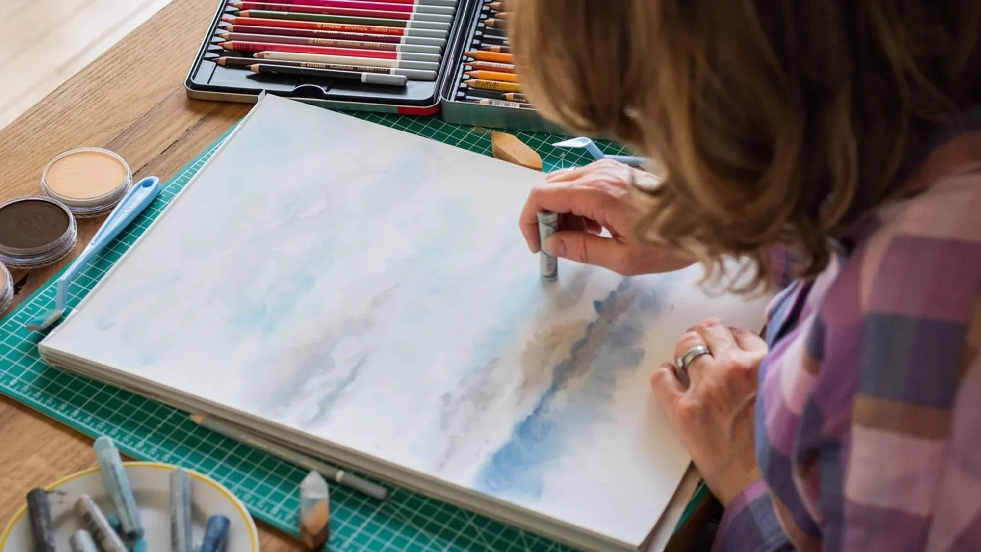 Clare Jones freelance illustrator based in Paris in her studio creating pastel drawing of the clouds, surrounded by art supplies including colored pencils, watercolors, and brushes on a wooden table.