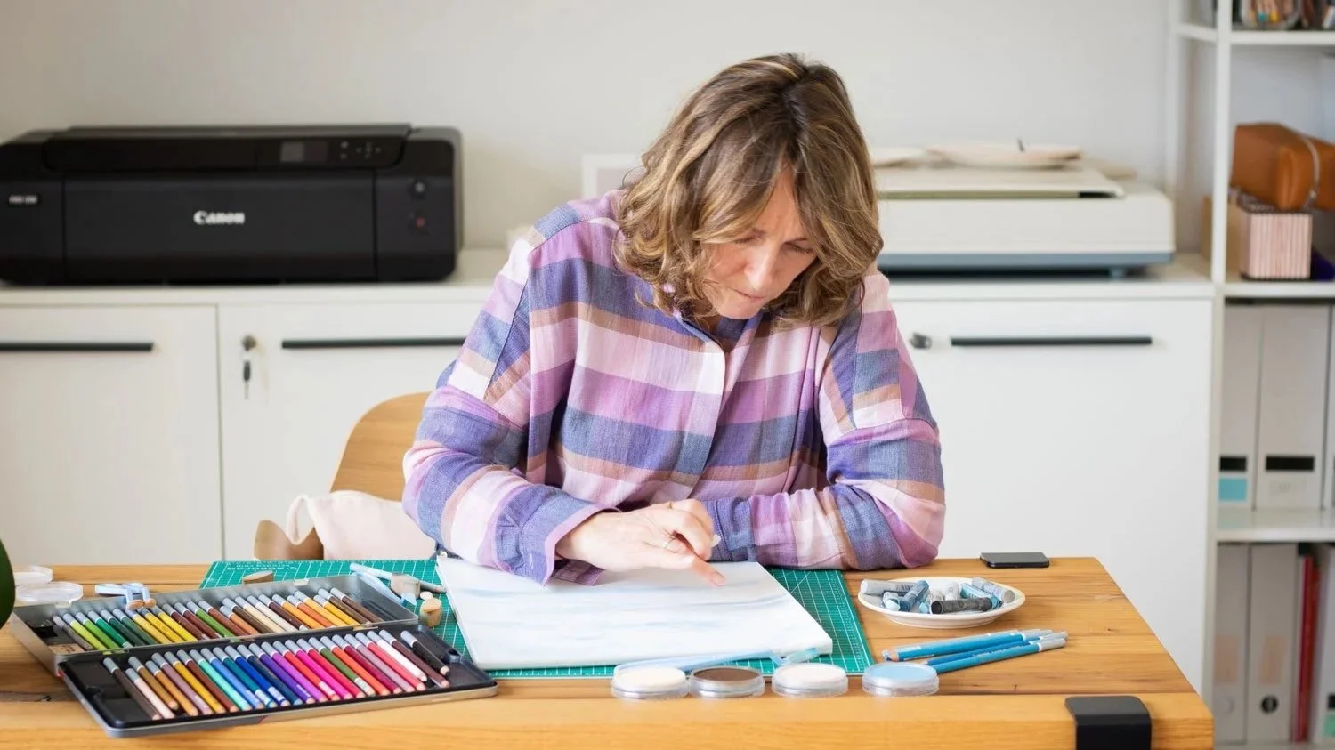Clare Jones freelance illustrator based in Paris working in her studio, surrounded by art supplies including colored pencils, chalks, and a notebook in a home or office setting.