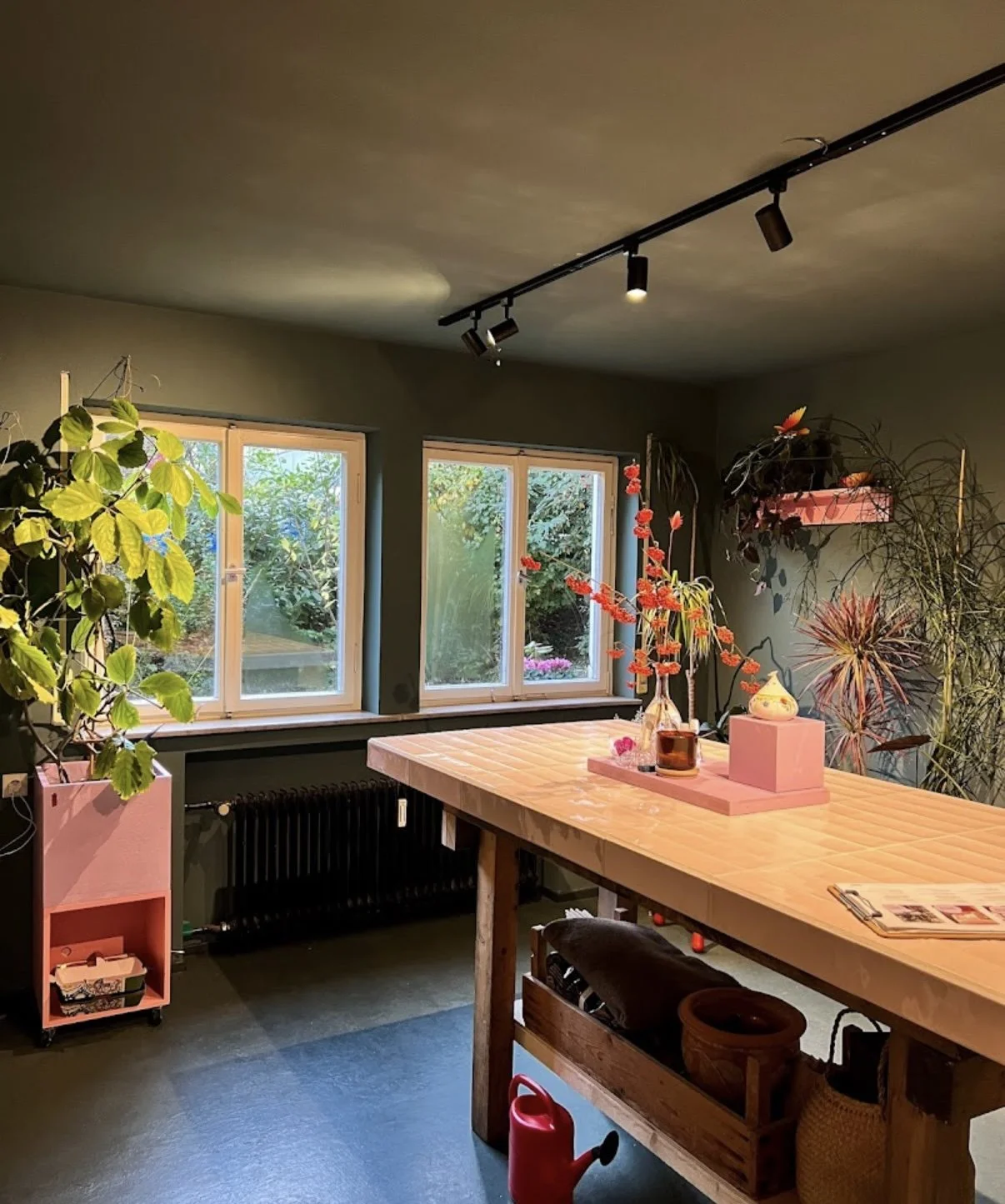 Interior of a room with three large windows, green walls, and a high wooden table decorated with flowers, candles, and magazines. There are several potted plants and a pink shelving unit.