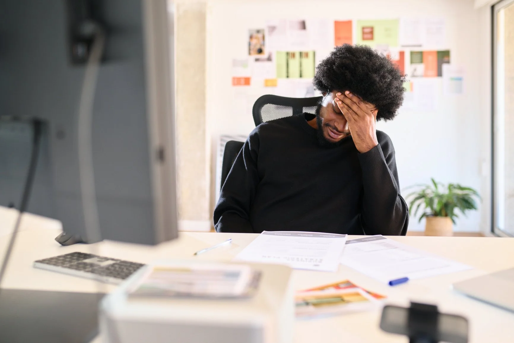 A man sitting at a desk with his hand on his forehead, appearing stressed or overwhelmed while working on papers in an office.