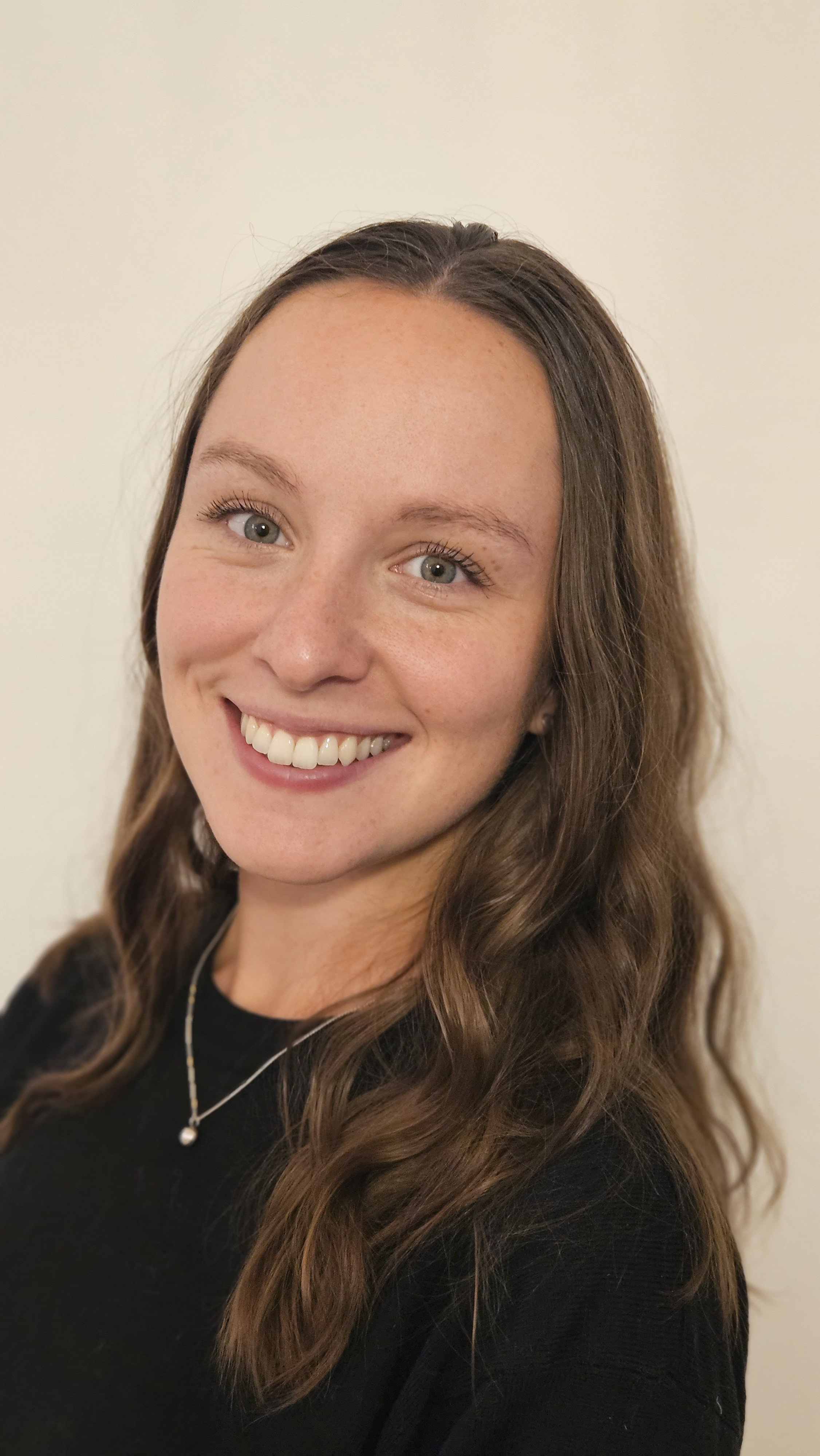 Portrait of a smiling young woman with long wavy brown hair, blue eyes, and a black top, against a plain light-colored background.