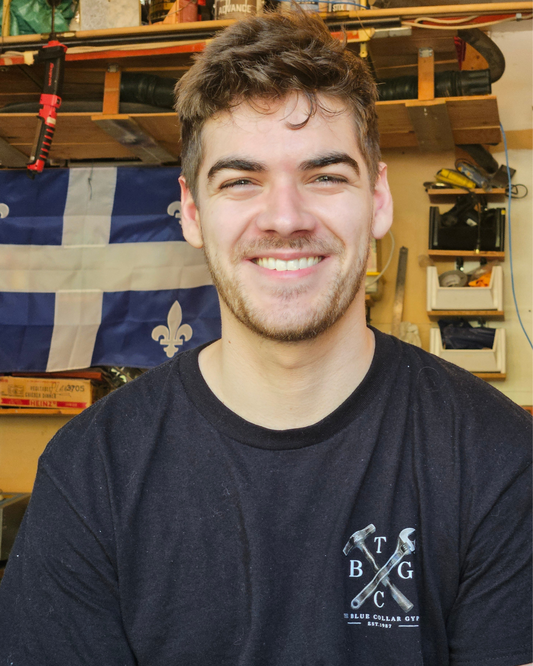 A young man with light brown hair and a beard, smiling in a workshop with tools and a Quebec flag in the background.
