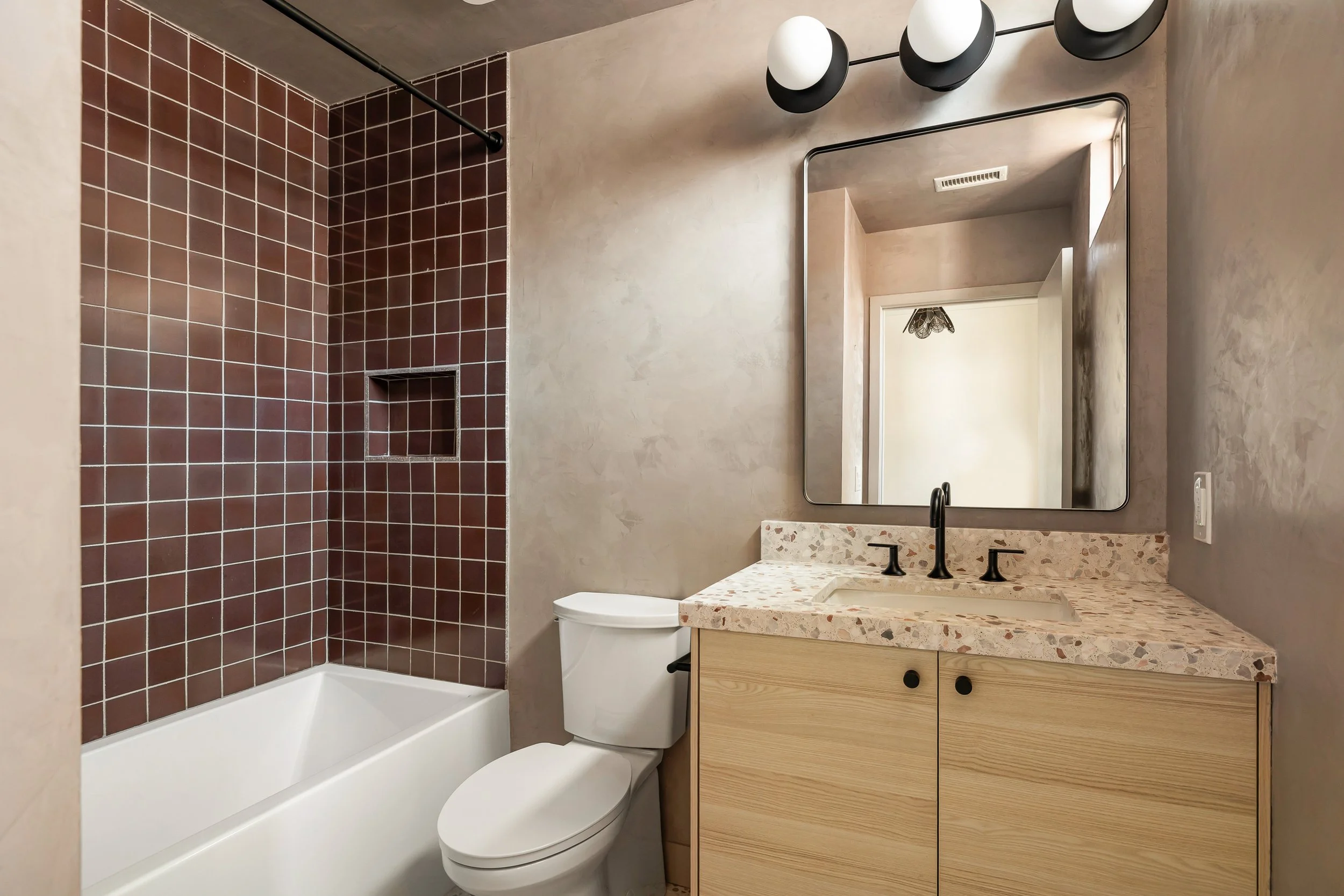 Modern bathroom with a bathtub, red tile shower area, white toilet, wooden vanity with a terrazzo countertop, black faucet, large mirror, and contemporary black and white wall-mounted light fixtures.