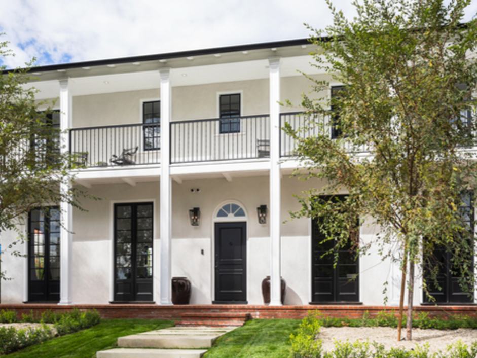 Front view of a modern two-story house with white exterior, black doors and windows, a balcony with black railing, and front yard with green lawn and trees.