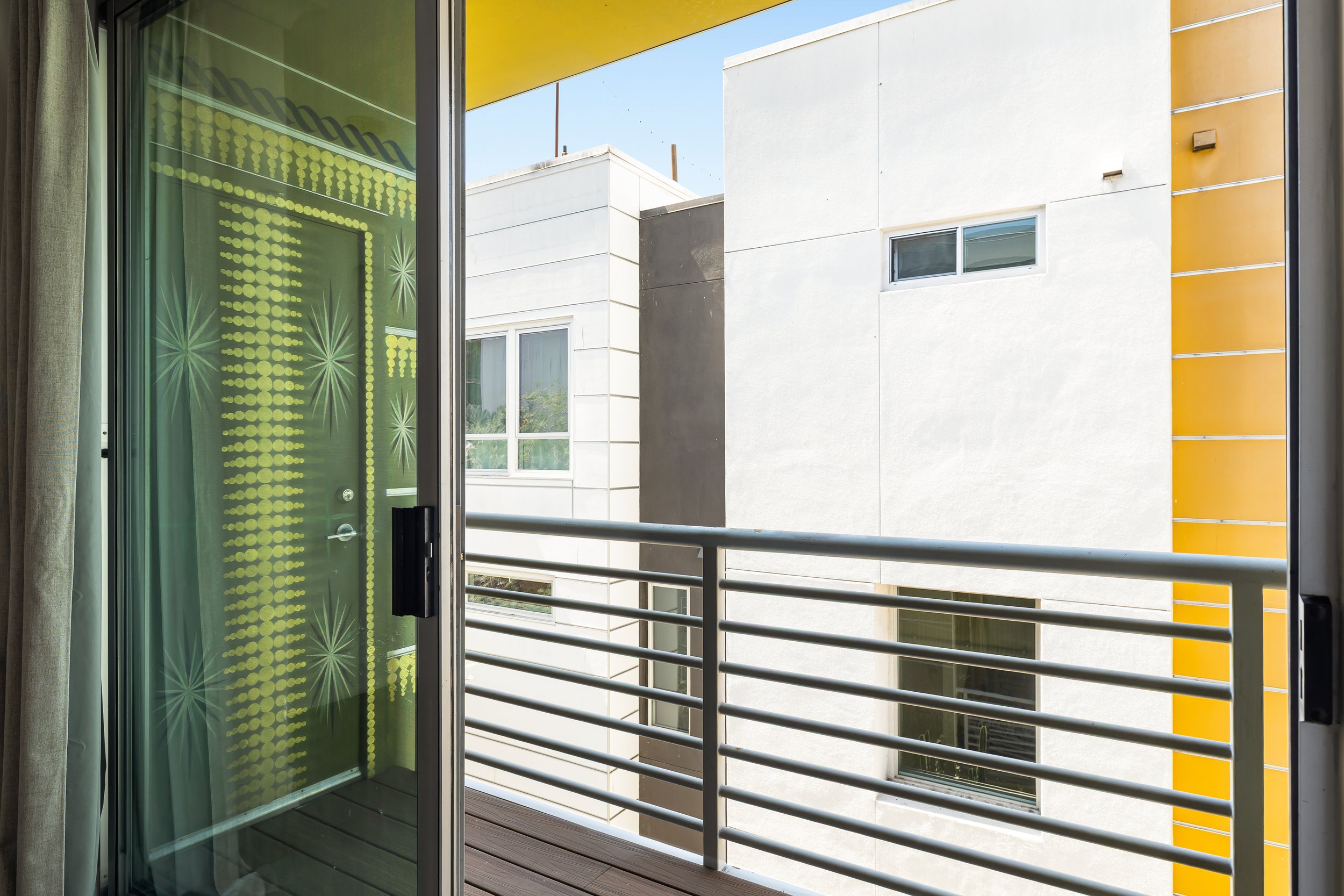 View from a sliding glass door opening to a balcony with metal railings. The balconies and building exterior are seen with white, gray, and yellow sections. There are windows on the building. The sky is clear and blue.