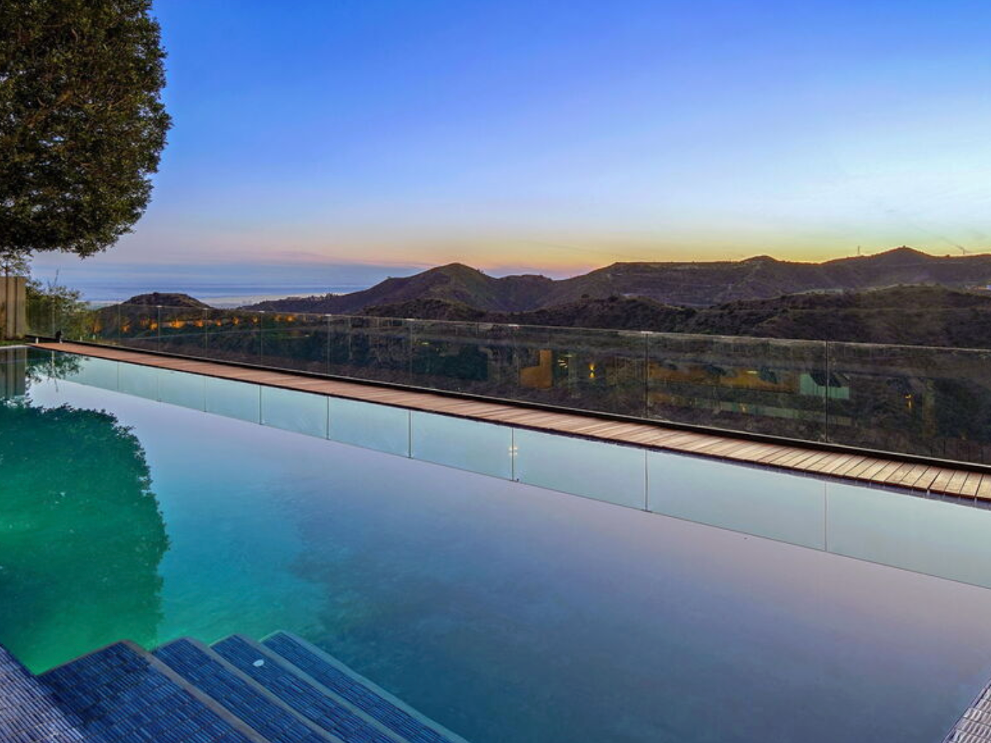 An infinity pool overlooking rolling hills and a distant ocean at sunset, with a glass fence surrounding the pool area.