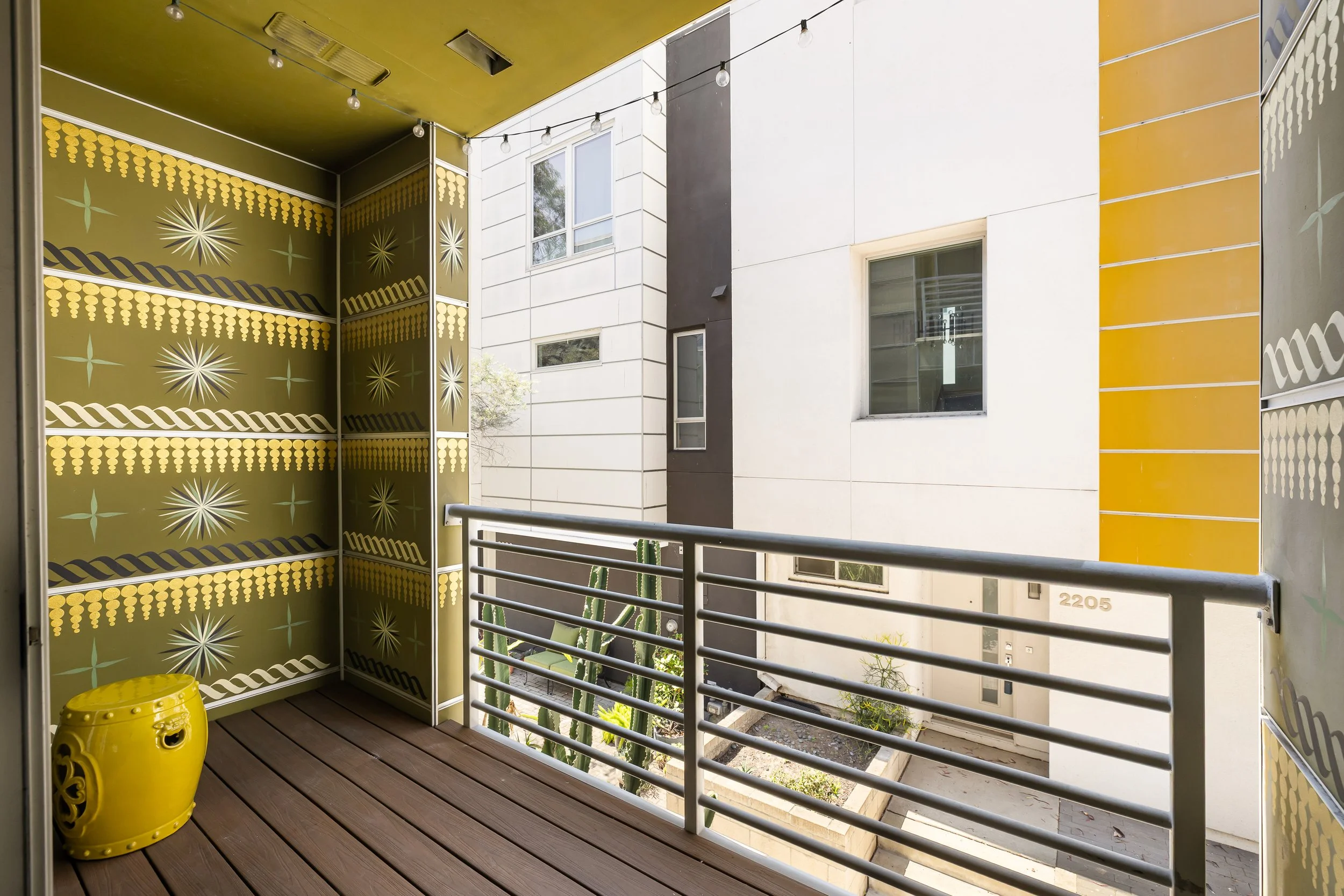 View of a small balcony with wooden deck flooring, a yellow ceramic stool, decorative yellow and black wall panels with patterns, a metal railing, two cactus plants, and a neighboring apartment building with windows and a door.