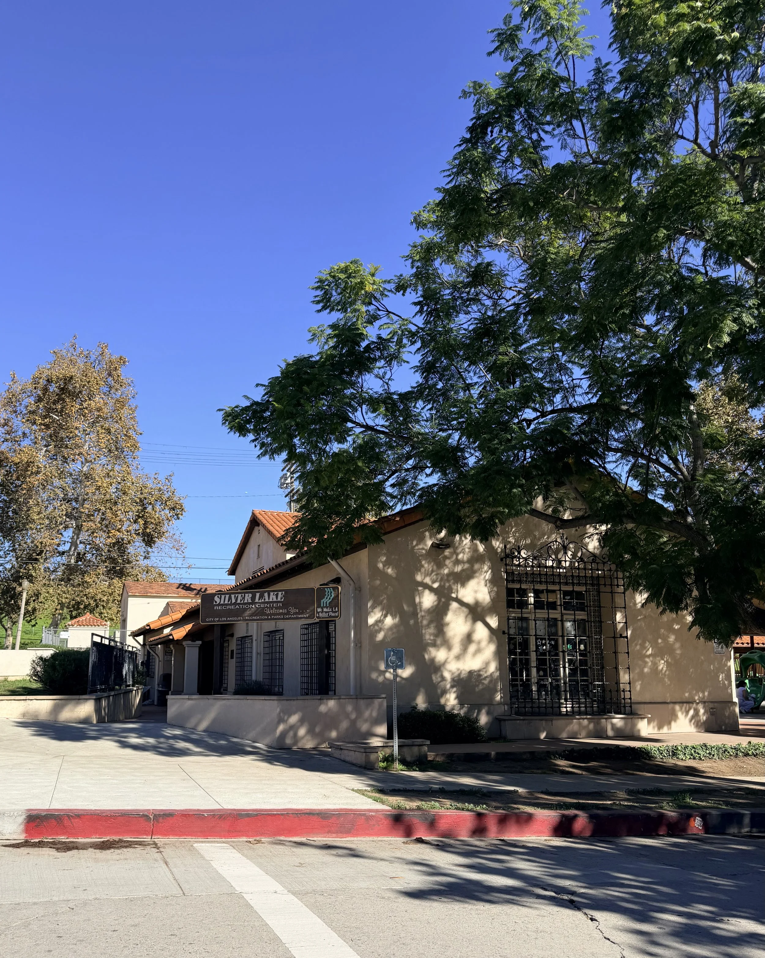 A beige building with a red tile roof, surrounded by trees, with a sign that reads 'Silver Lake Recreation Center,' in front of a sidewalk and street.