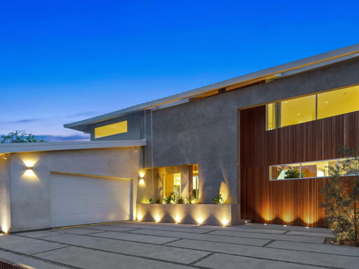 Modern house exterior at twilight, with well-lit driveway and large windows revealing interior lighting and greenery inside.