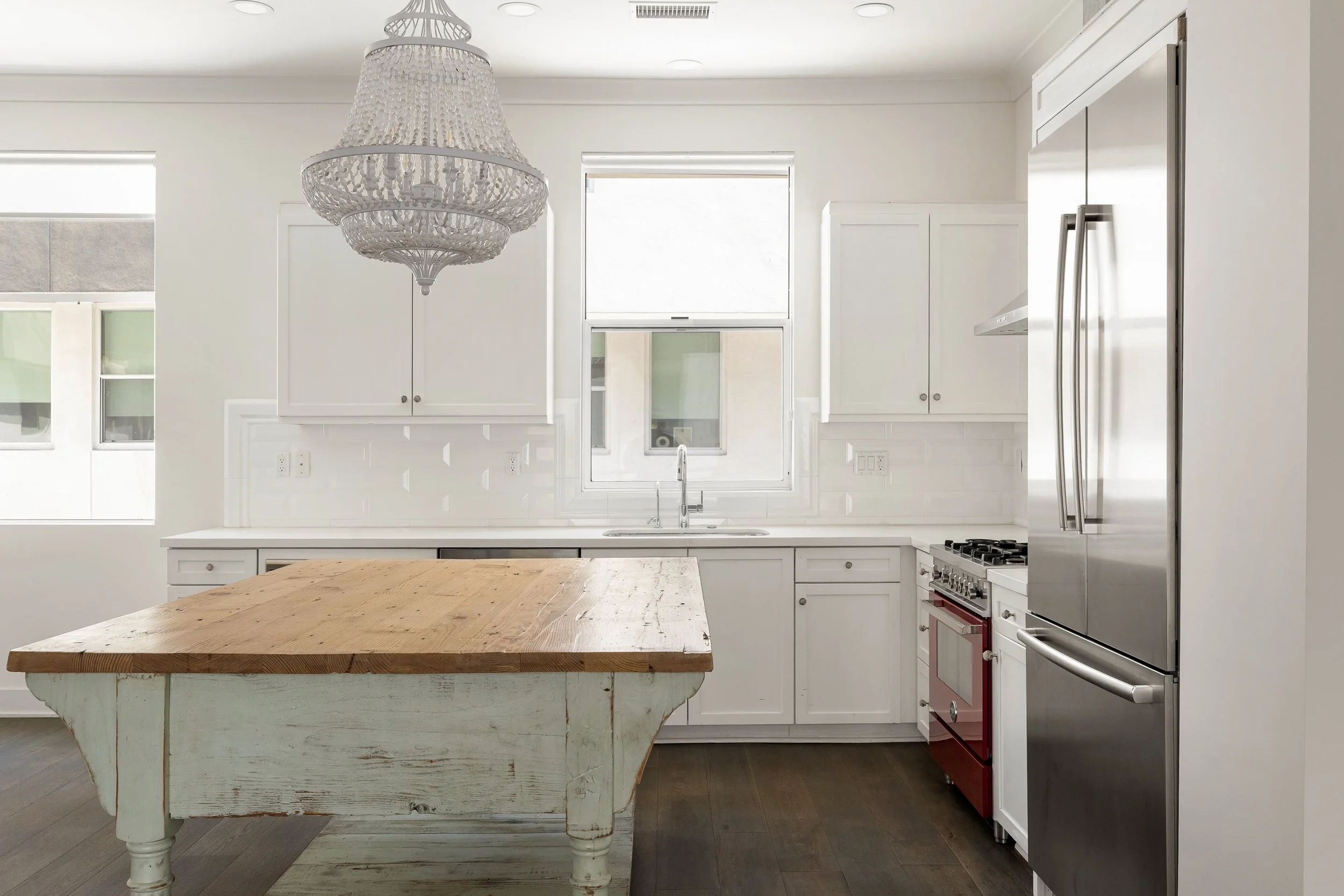 A bright, modern kitchen featuring white cabinets, a large wooden kitchen island, a stainless steel refrigerator, and a classic red and white oven. There is a decorative chandelier hanging from the ceiling and two windows above the sink letting in natural light.