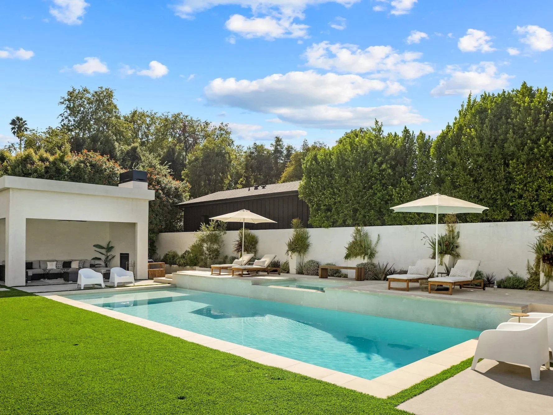 Modern backyard with a swimming pool, lounge chairs, umbrellas, and green landscaping under a partly cloudy sky.