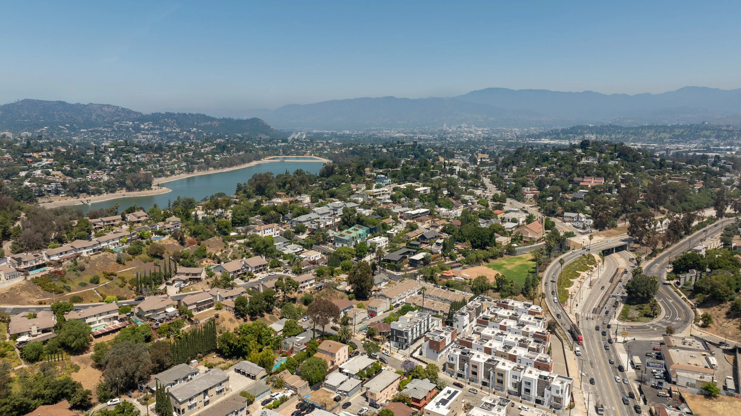 Aerial view of a city with a river flowing through residential neighborhoods, surrounded by hills and mountains under a clear blue sky.