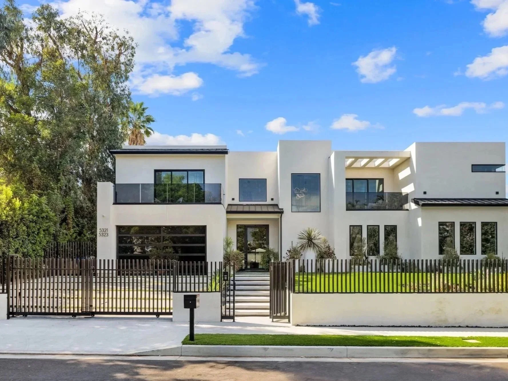 Modern white multi-story house with flat roof, large windows, and a black metal gate at the entrance. The house has a small front yard with green grass, some shrubs, and a pathway leading to the front door. Tall trees and a blue sky with a few clouds are visible in the background.